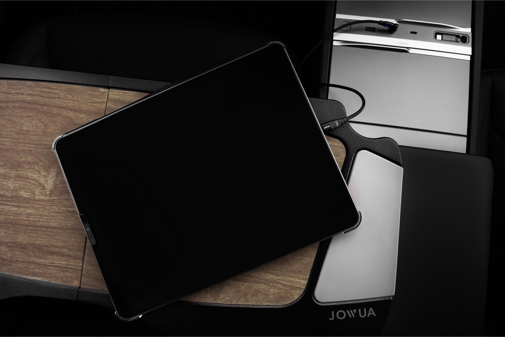 A black-screen tablet connects to a Tesla center console via cable amid wood and metal panels featuring control buttons. Partially covering the armrest, etched with "JOWUA," it provides an elegant charging/storage solution. Compatible with Tesla Model Y Juniper 2025+ and Model 3 2024+.