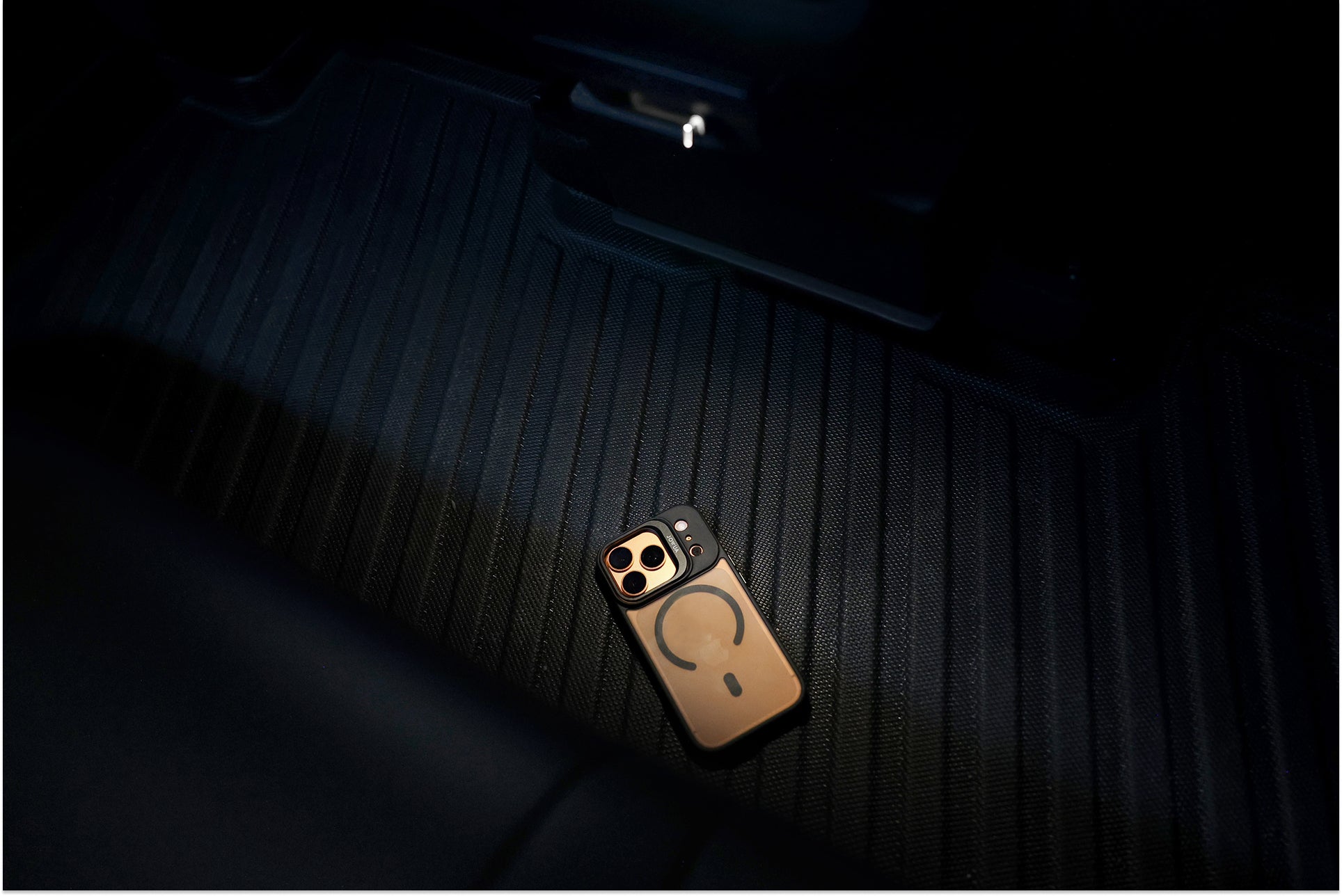 A gold smartphone with a MagSafe ring on its case lies face down on the black vehicle floor mat, partially illuminated by the JOWUA 130W LED Rear Vent Hub, custom-fit for Model 3 Highland 2024+ and Model Y Juniper 2025+.