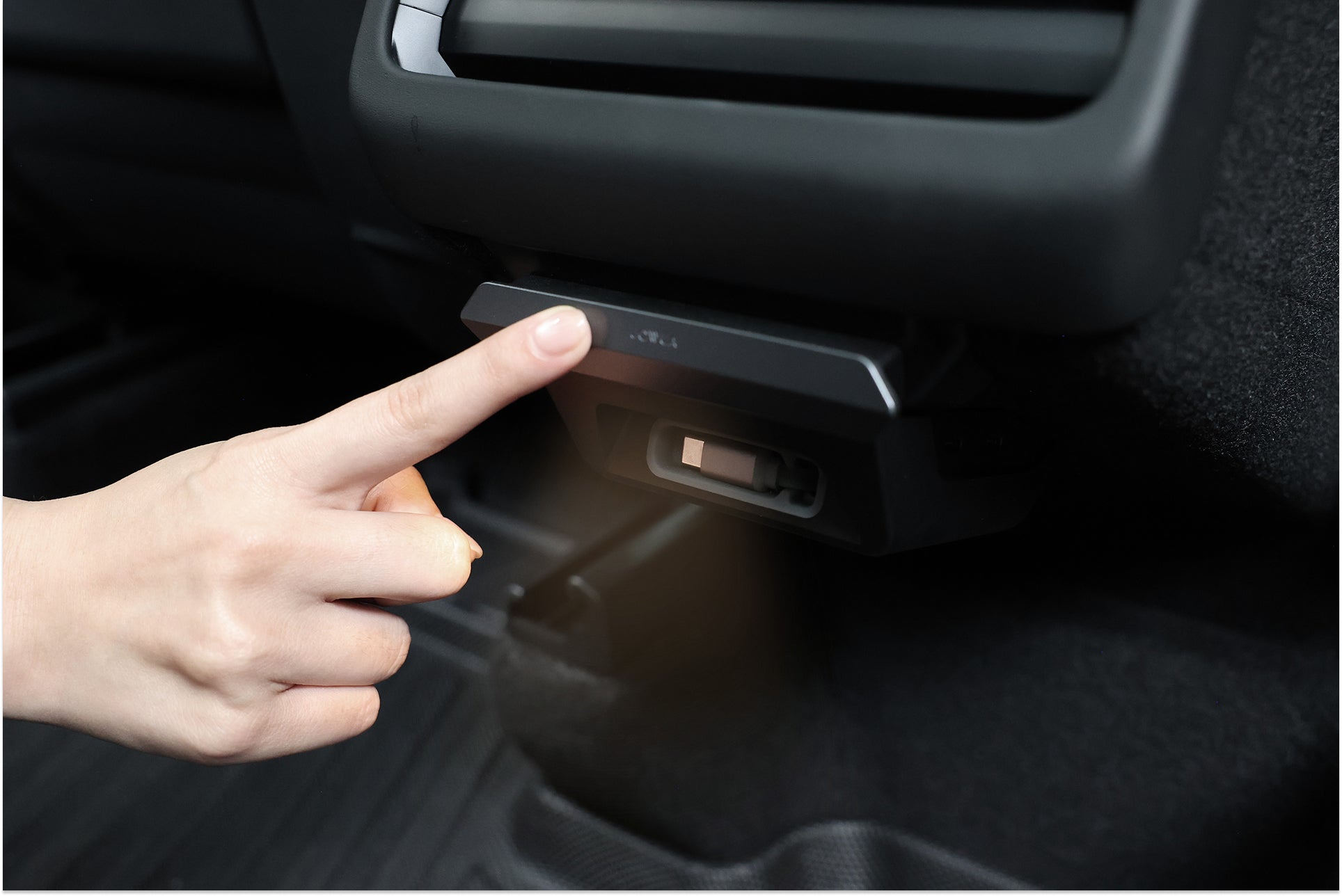 A hand presses the rectangular JOWUA 130W LED Rear Vent Hub for Model 3 Highland 2026+ / Model Y Juniper 2026+, located below the car’s rear seat air vent. The interior has dark upholstery and a plastic panel.