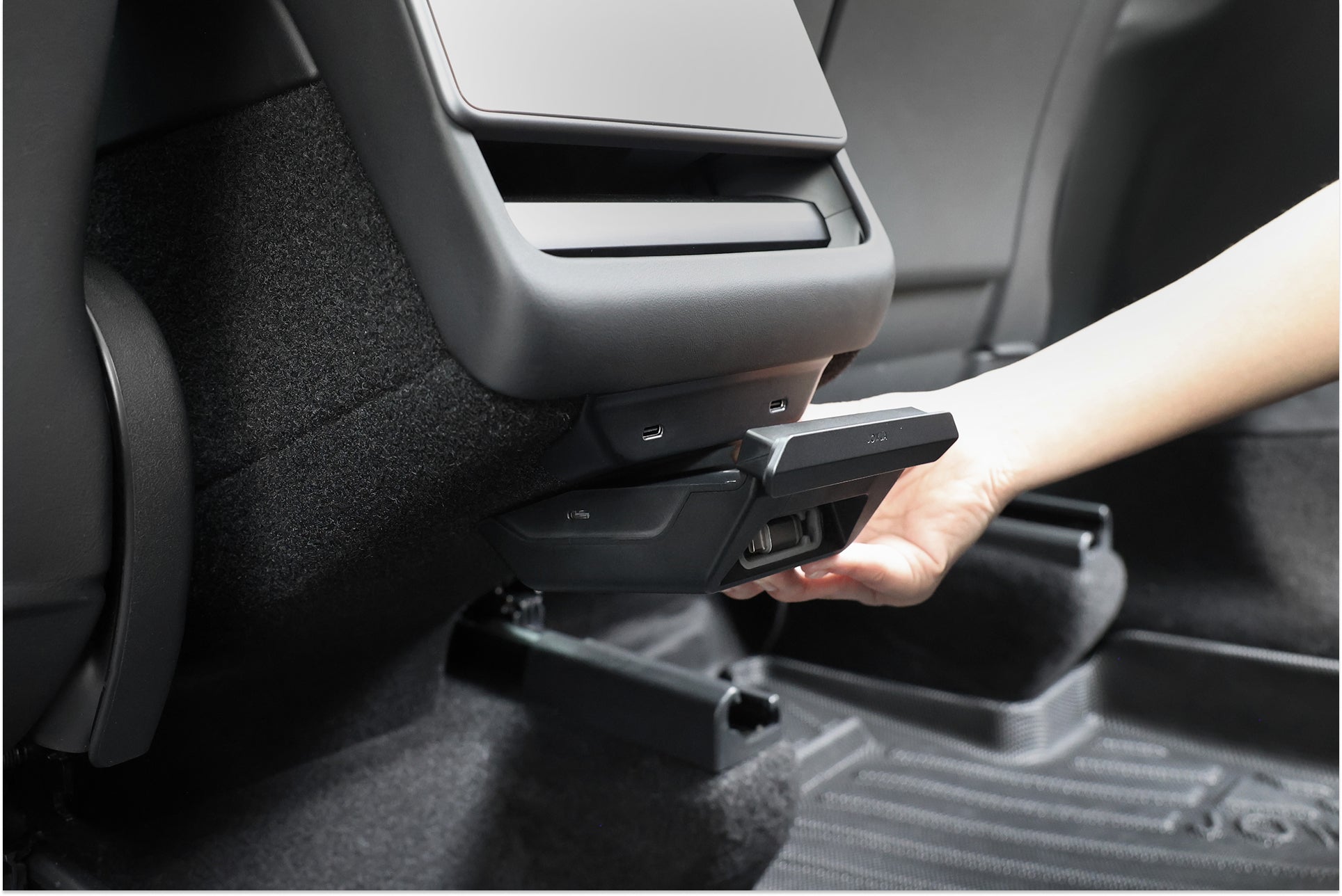 A hand pulls down a JOWUA 130W LED Rear Vent Hub (Model 3 Highland 2024+/Model Y Juniper 2025+) panel behind the center console, with black carpeting and floor mats visible inside the car.