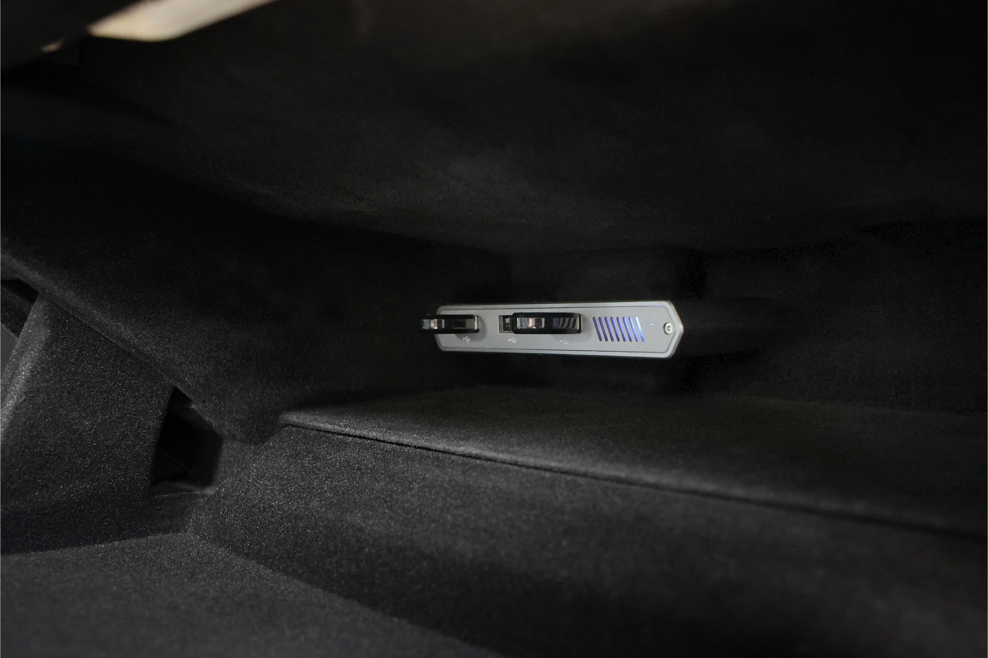 A close-up of the JOWUA Tesla 1TB SSD + 3-Port HUB, an electronic device with buttons and vents, mounted inside a dark carpeted compartment—likely a car's glove box for expanded Tesla Sentry Mode storage.