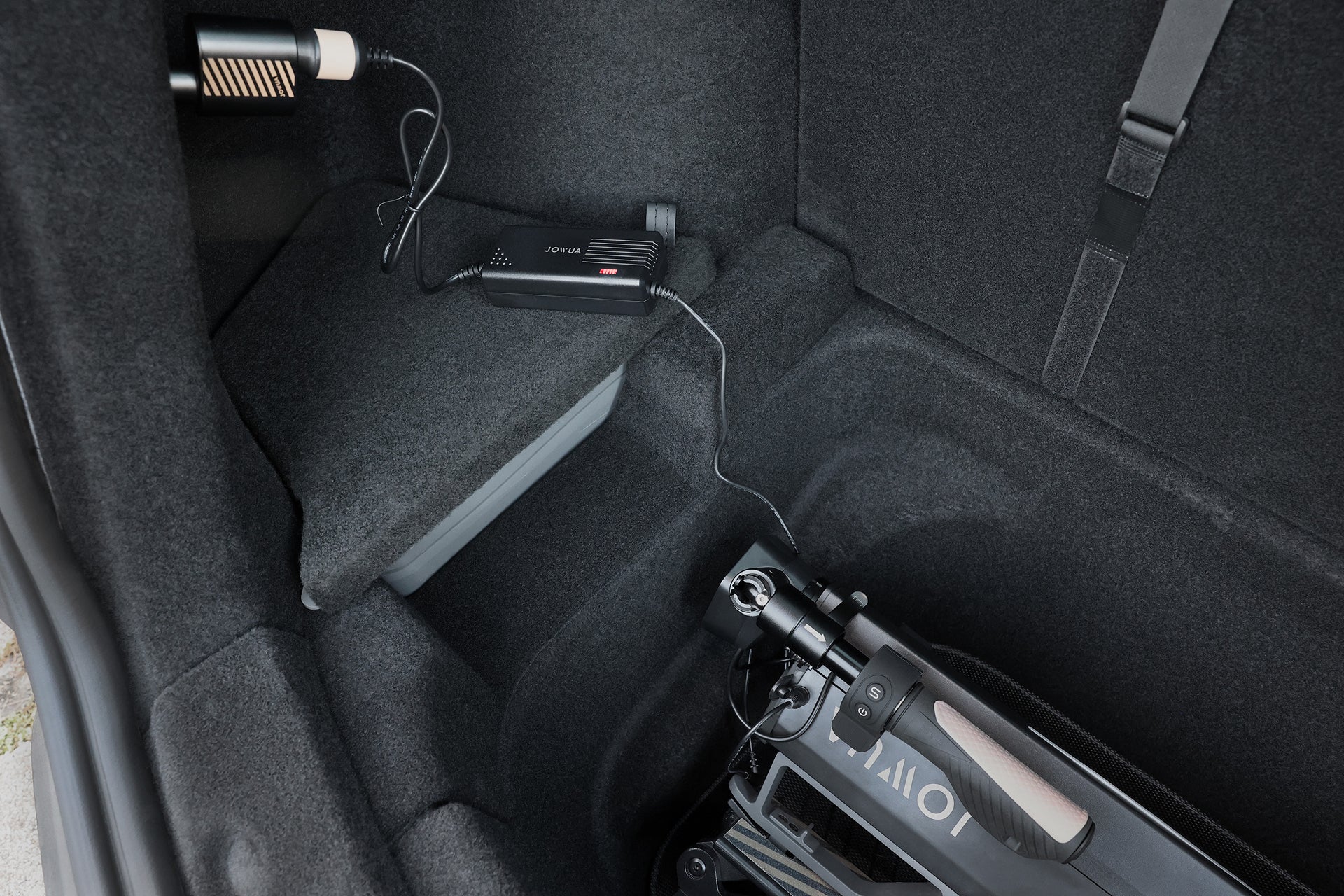 A black electric scooter is stored in a car trunk as a JOWUA 3 in 1 Type-C Car Charger, plugged into a socket, charges the scooter with visible cables against the dark interior lining.