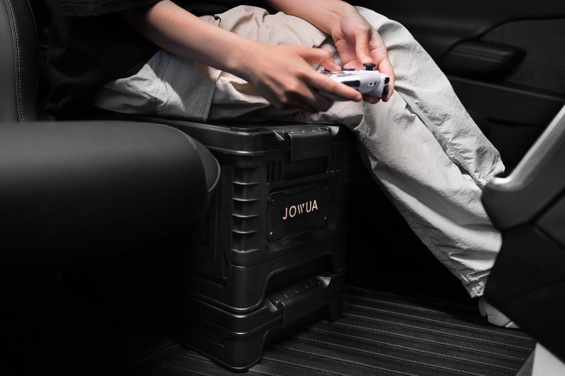 Inside the vehicle, a person holds a gaming controller, their hands resting on a sturdy 30L Stackable Storage Box and Tesla Trunk Organizer with the "JOWUA" brand. They