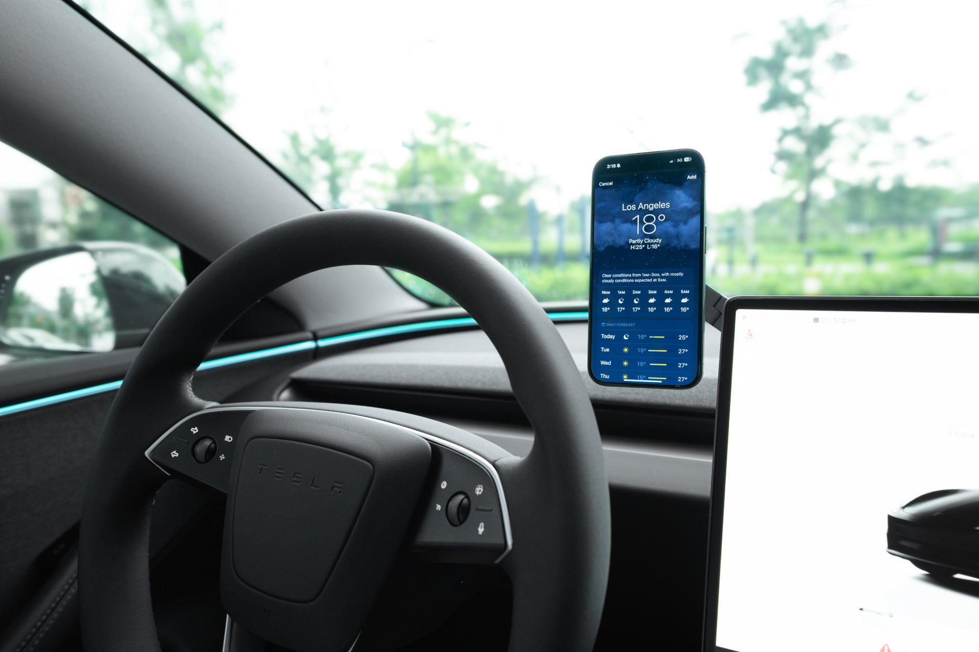 A Tesla steering wheel and dashboard are shown. A JOWUA 6D Tesla Compatible Invisible Foldaway Car Mount Qi2 Wireless Charger (MagSafe) holds a smartphone beside the wheel, displaying a Los Angeles weather app. Part of the digital screen is visible on the right.