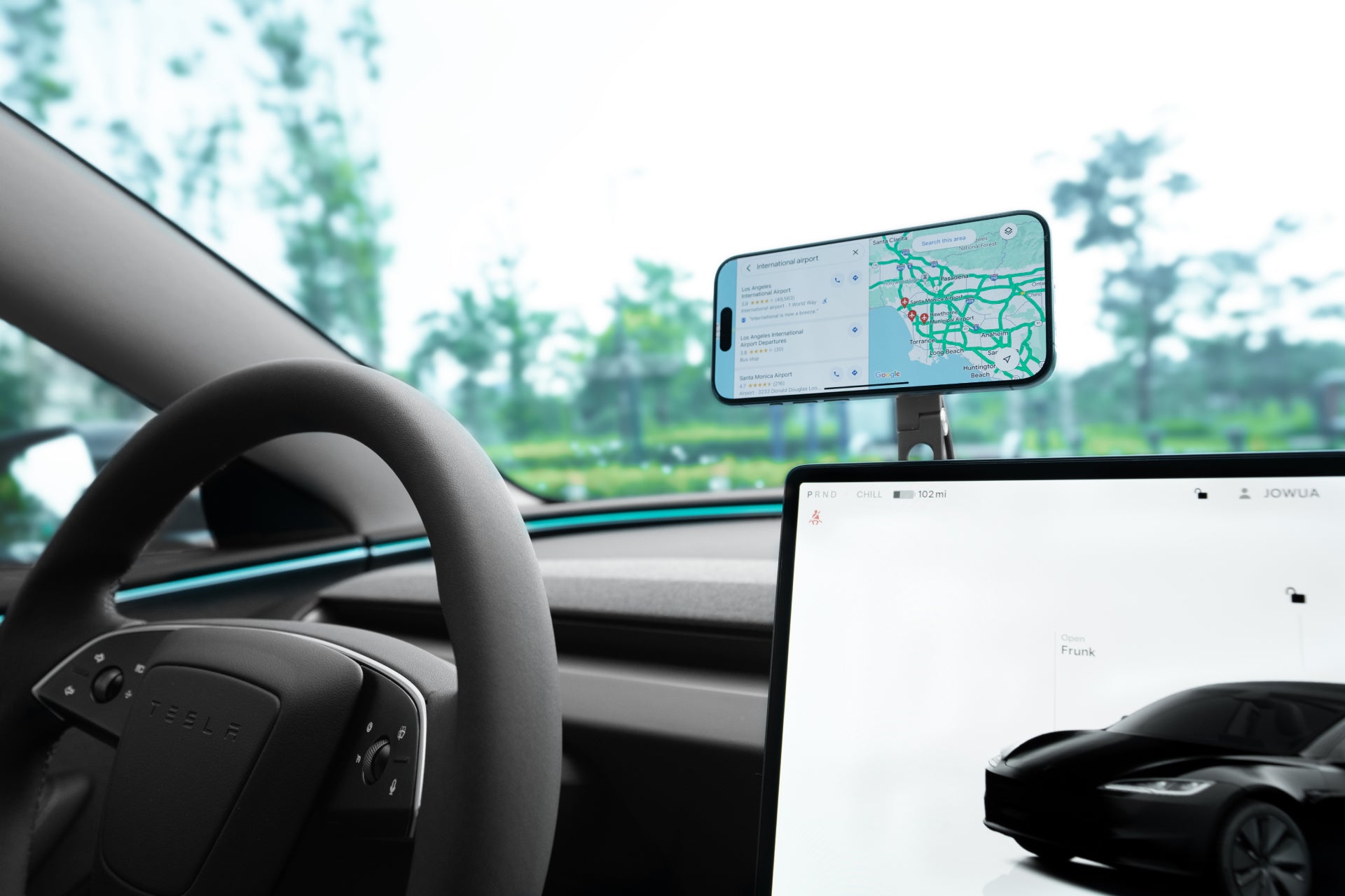 A Tesla interior shows a JOWUA 6D Invisible Foldaway Car Mount Qi2 Wireless Charger (MagSafe) holding a smartphone, with a steering wheel, dashboard screen, and greenery plus a blurred city view visible through the windows.