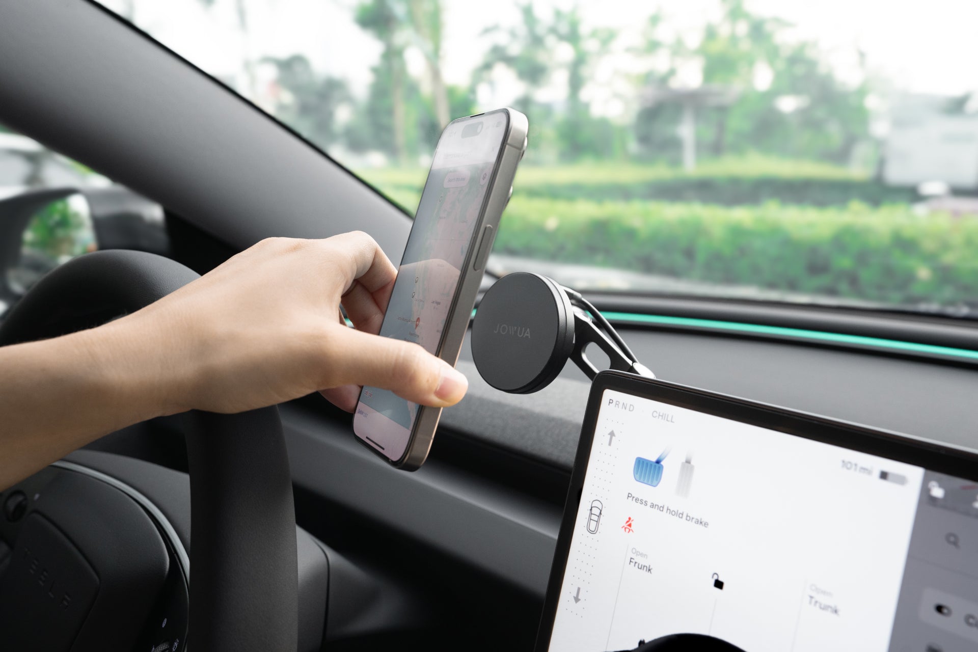 A person places a smartphone onto the JOWUA 6D Tesla Compatible Invisible Foldaway Car Mount Qi2 Wireless Charger (MagSafe) near the dashboard, with car controls on display and greenery visible outside the window.