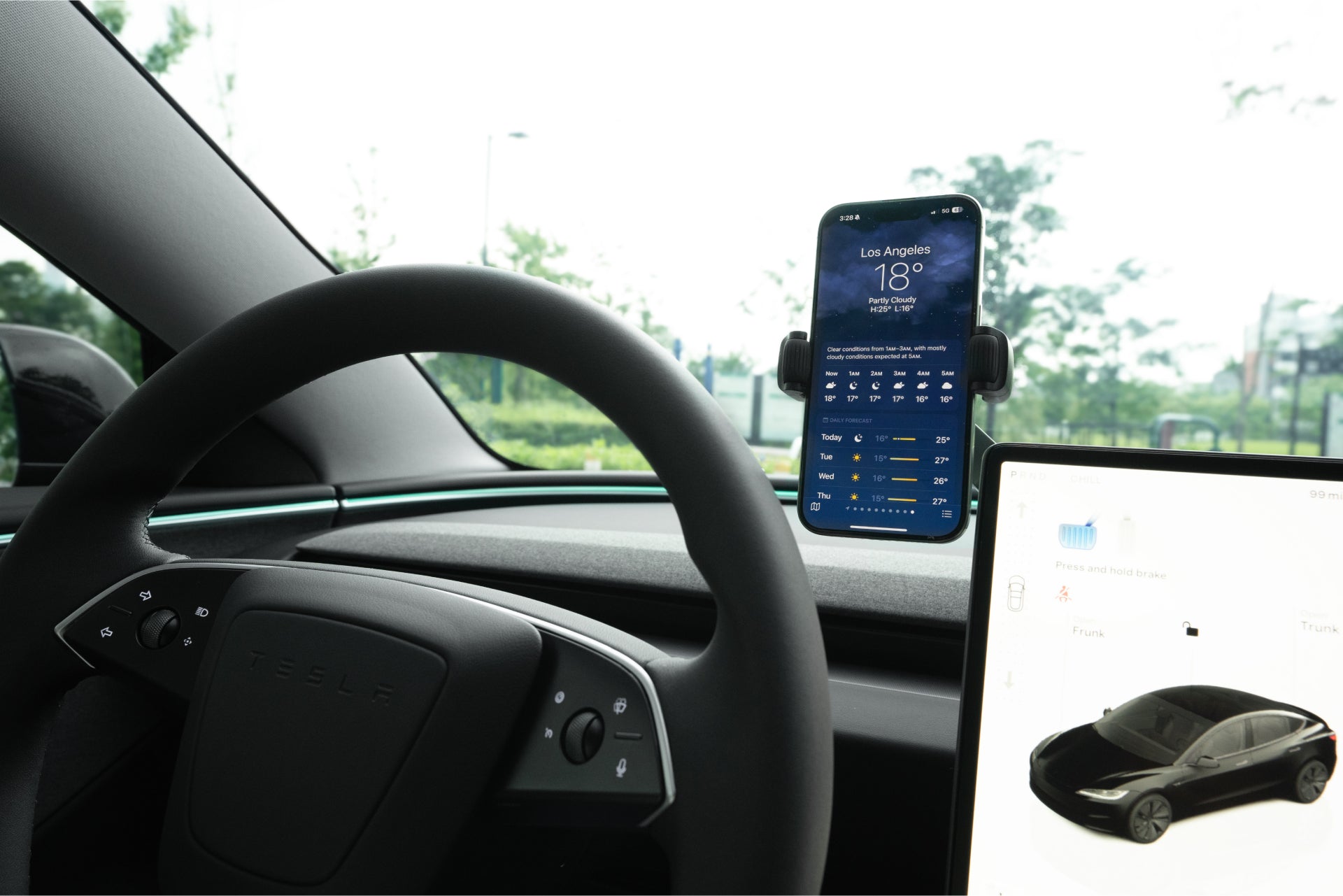 A JOWUA 6D Tesla Compatible Invisible Foldaway Car Mount Qi2 Wireless Charger holds a smartphone showing a weather app on the dashboard near the steering wheel and touchscreen, with green trees visible through the Tesla’s windshield.