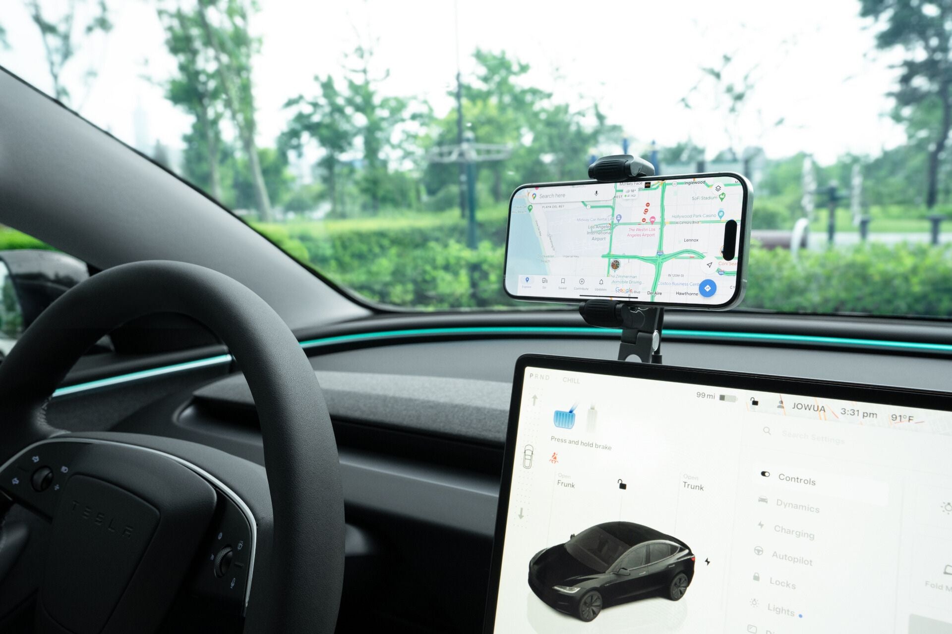 A smartphone with a navigation app rests on the JOWUA 6D Tesla Compatible Invisible Foldaway Car Mount Qi2 Wireless Charger, mounted by the touchscreen’s vehicle controls. Trees and greenery are visible outside the windshield.