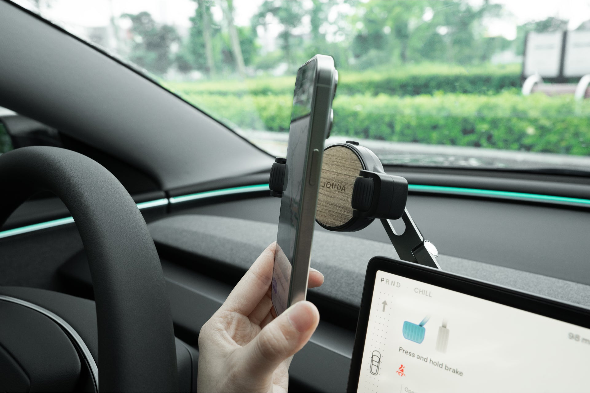 A person mounts a smartphone onto the JOWUA 6D Tesla Compatible Invisible Foldaway Car Mount Qi2 Wireless Charger, with the steering wheel and digital climate controls visible, showcasing convenient MagSafe charging for Tesla S/3/X/Y/Cybertruck.