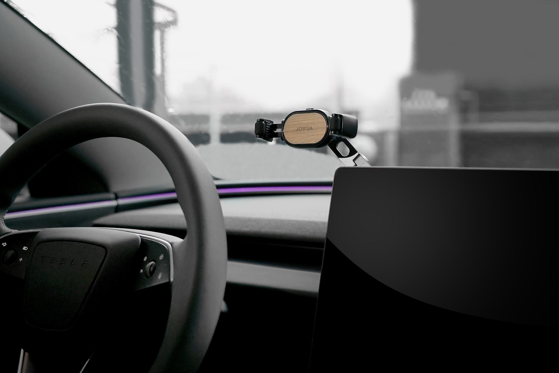 Inside the car, a steering wheel is on the left, while a JOWUA 6D Invisible Foldaway Car Mount sits innovatively on the right with a stylish finish. The foggy windshield reveals urban architecture, indicative of refined design elements and adjustable angles for optimal viewing.