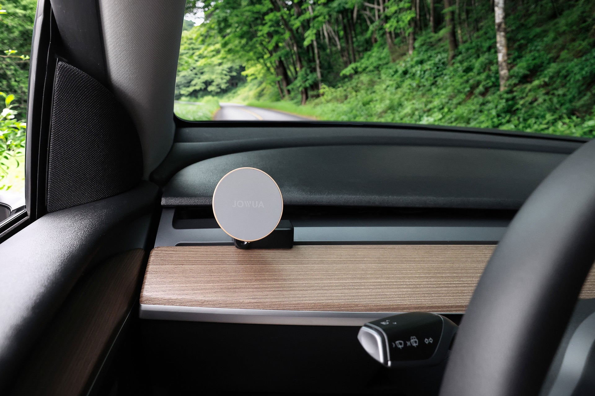 The JOWUA Active Cooling MagSafe Car Mount for Air Vents (Model 3/Y 2018-2024) is installed on a wood-trim air vent next to the steering wheel, in a parked car beside a lush green forest road.