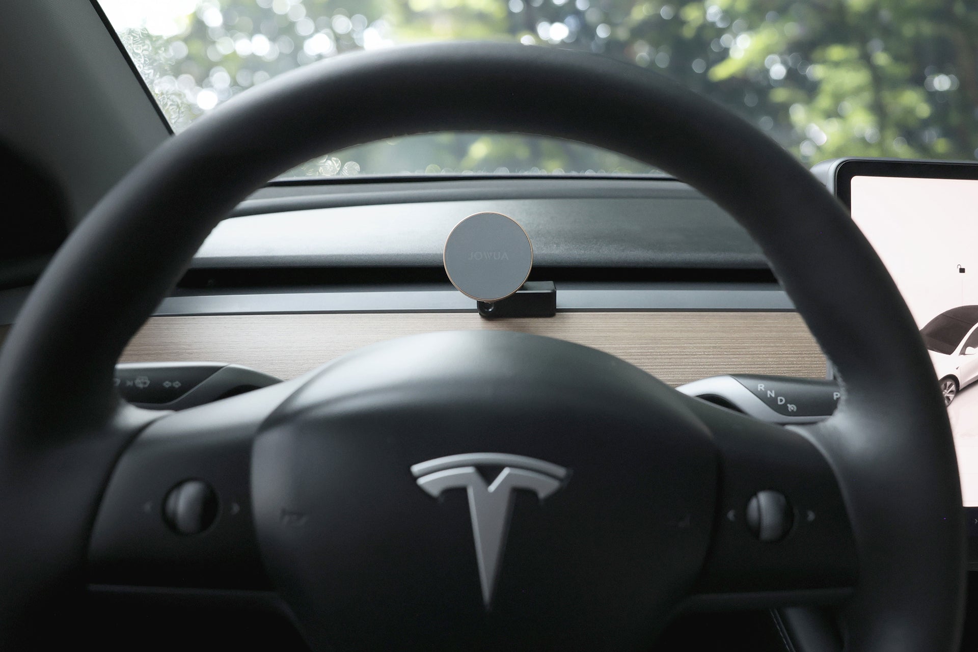 View from the driver’s seat of a Tesla, featuring the Tesla logo on the steering wheel, a wooden dashboard, and a JOWUA Active Cooling MagSafe Car Mount for Air Vents (Model 3/Y 2018-2024), with greenery visible through the windshield.