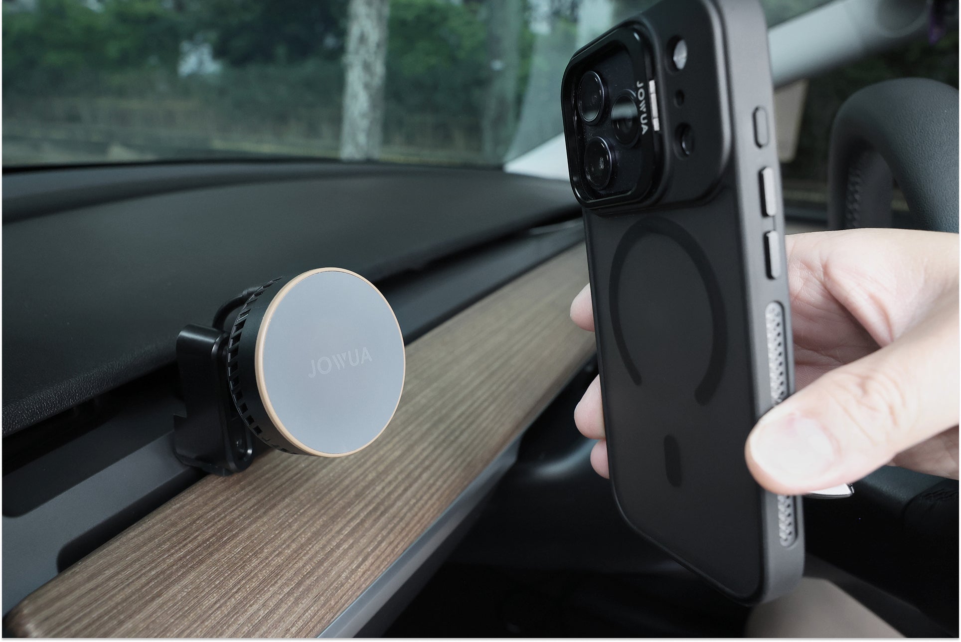 A hand holds a smartphone with a magnetic case near a JOWUA Active Cooling MagSafe Car Mount for Air Vents (Model 3/Y 2018-2024) attached to a car dashboard with wood trim.