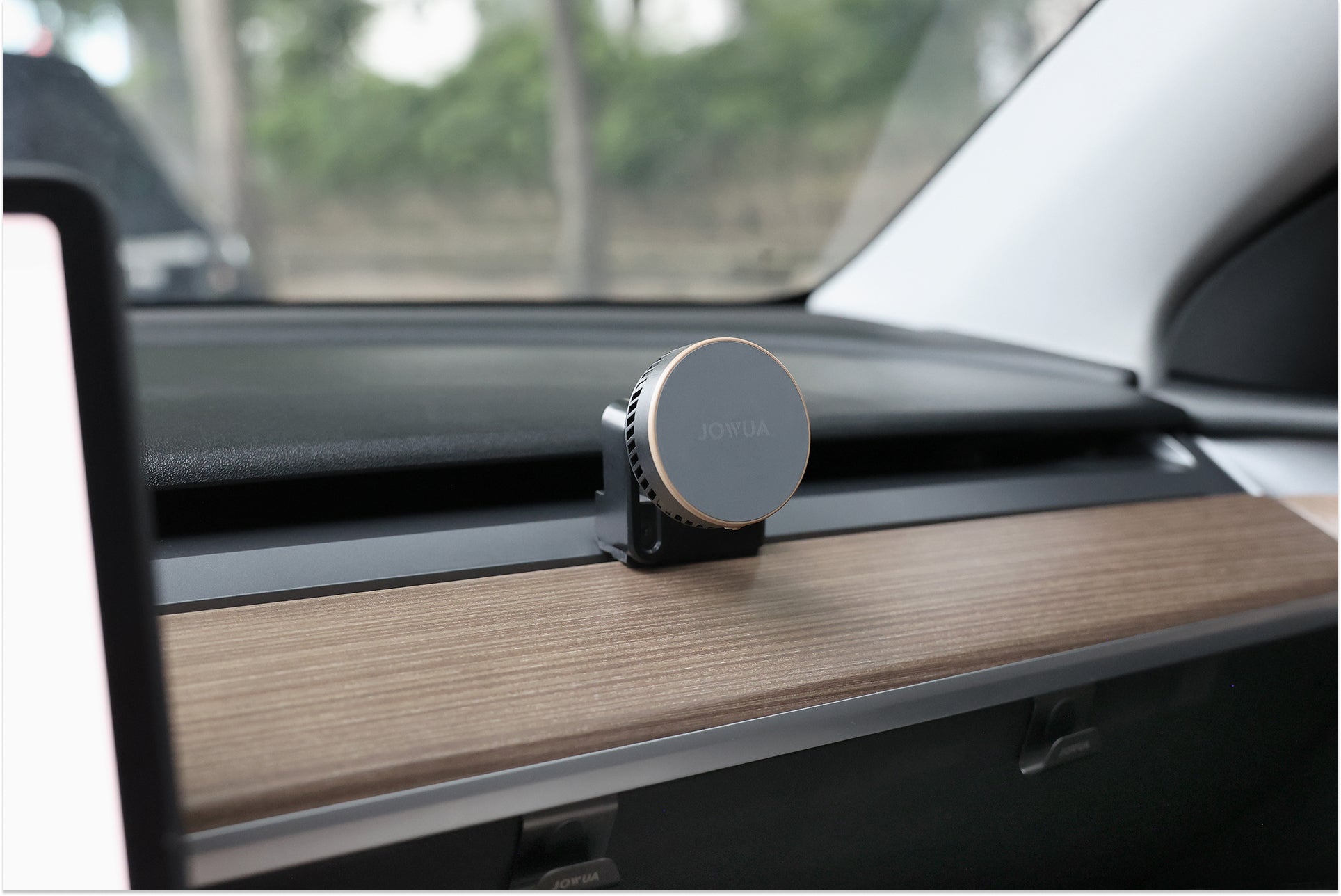 A JOWUA Active Cooling MagSafe Car Mount for Air Vents (Model 3/Y 2018-2024) is attached to a car’s wooden dashboard trim above the glove compartment, with trees visible outside the window—compatible with Model 3/Y.