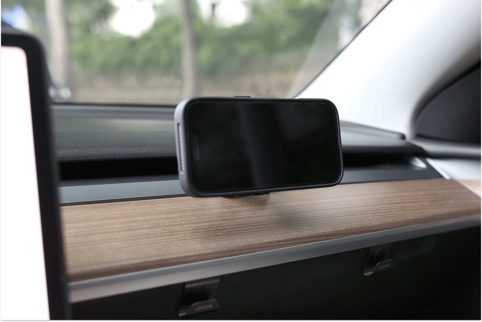A JOWUA Active Cooling MagSafe Car Mount for Air Vents (Model 3/Y 2018-2024) holds a smartphone in a black case, mounted horizontally on a wooden dashboard trim with trees and greenery visible through the car windshield.