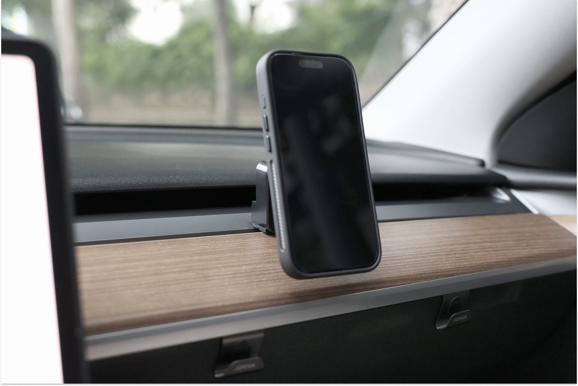A JOWUA Active Cooling MagSafe Car Mount for Air Vents (Model 3/Y 2018-2024) secures a black-cased smartphone to the wooden dashboard trim, with trees and greenery visible outside the car window.