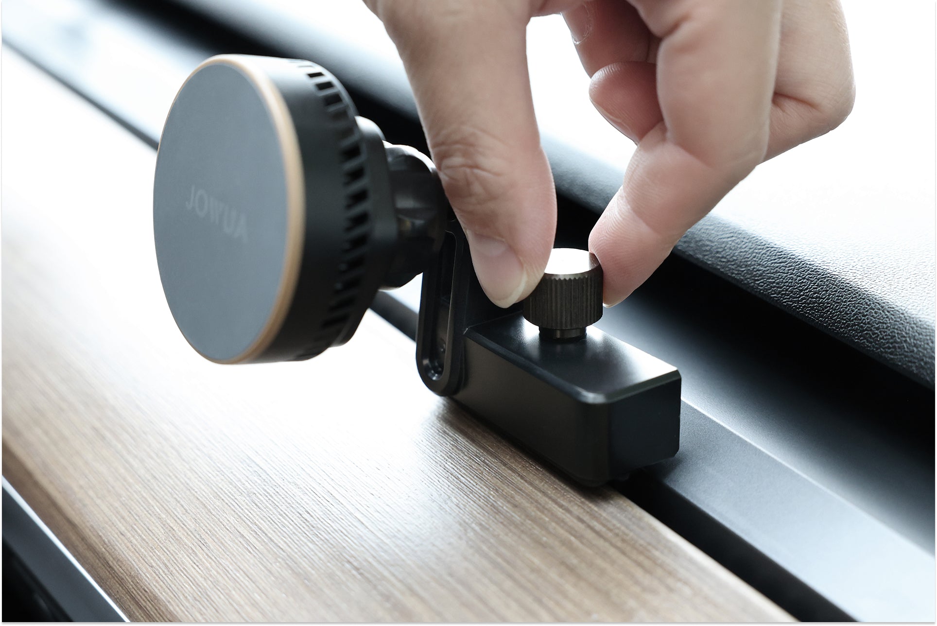 A close-up of a hand tightening a knob on the JOWUA Active Cooling MagSafe Car Mount for Air Vents (Model 3/Y 2018-2024), securely attached to a wooden and metal surface—perfect for holding your phone or charging with Qi2 fast charging.