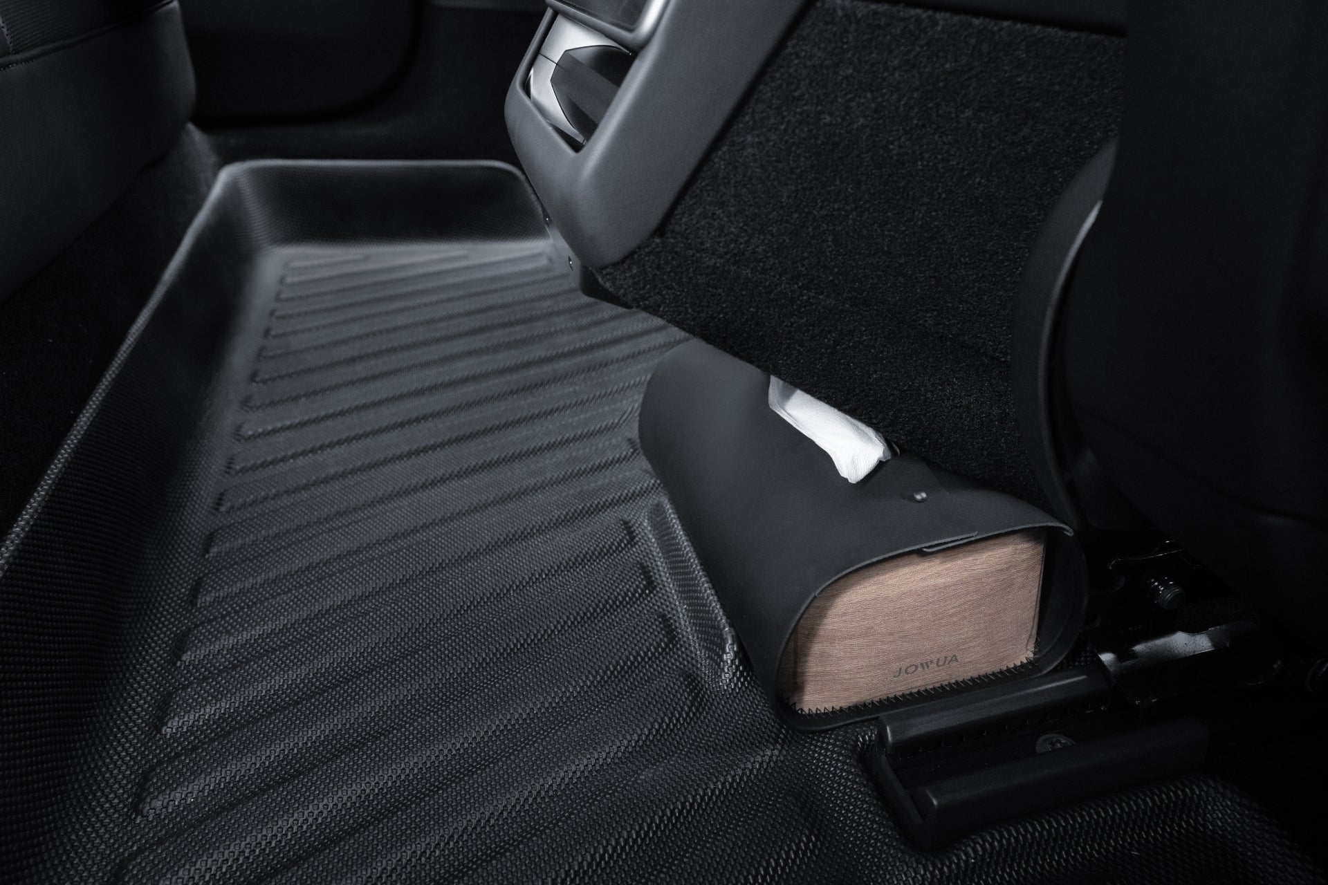 Close-up of a car's interior focusing on the backseat area. A JOWUA Model 3 All-Weather Floor Liner with a textured black design covers the floor, showcasing its custom fit. A box with a partially exposed tissue is placed against the back of a seat, adding a functional touch to the clean, minimalist setting.