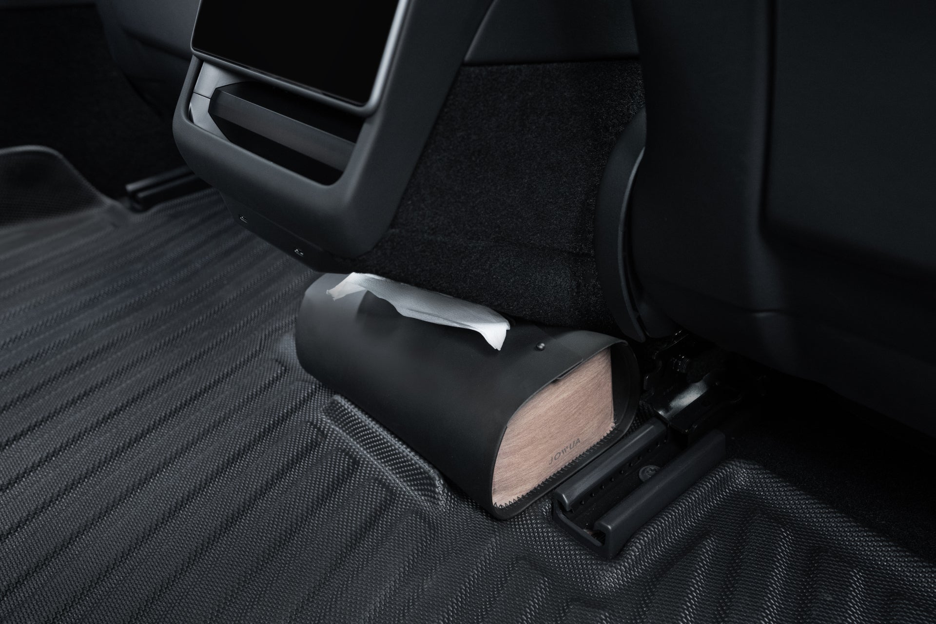 A wooden block wedge secures a car seat on JOWUA's Model 3 All-Weather Floor Liners, with a white paper partially sticking out from the block's cover. The dark, modern interior complements these floor liners, providing both style and functionality for the Tesla Model 3.