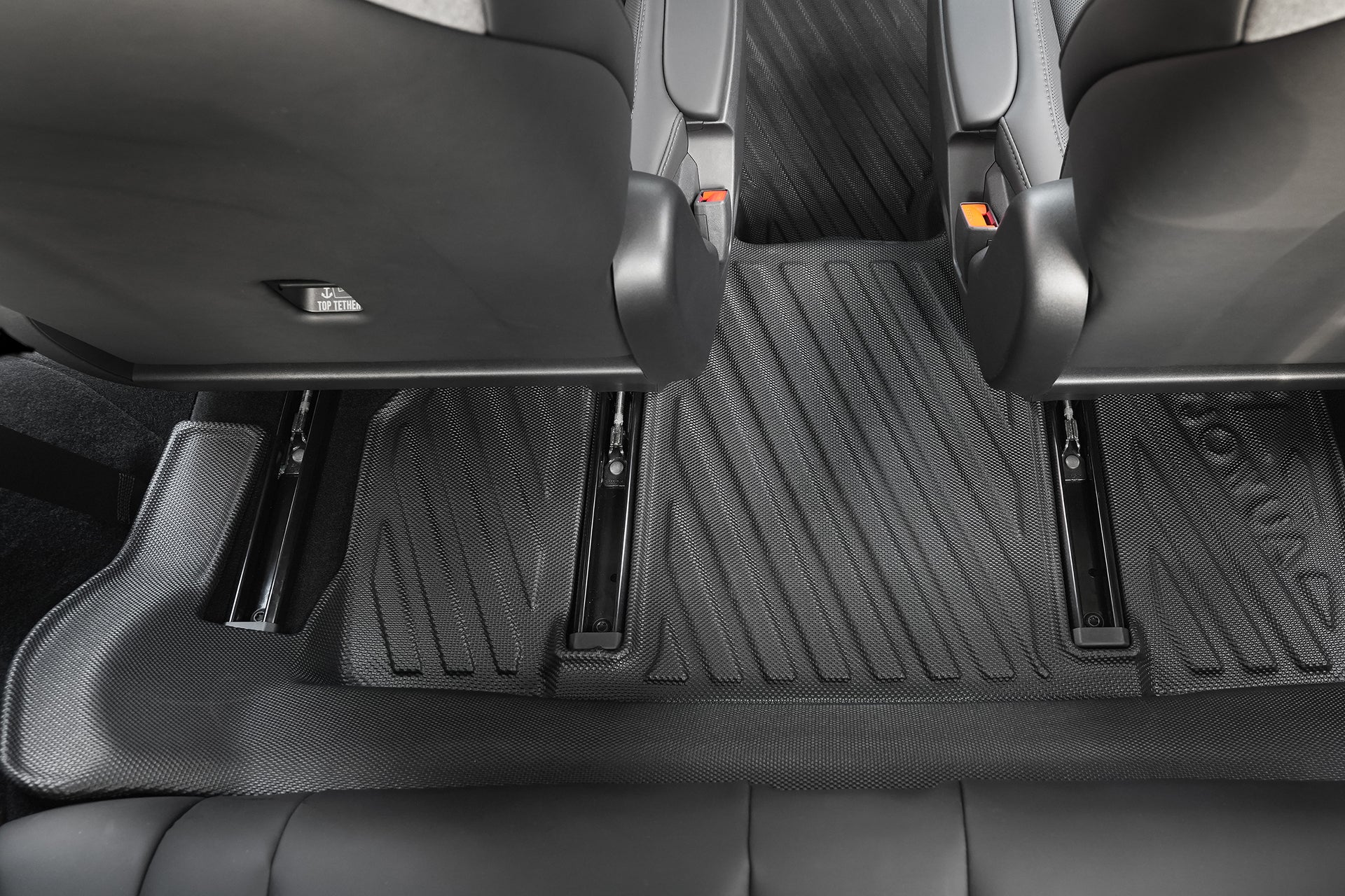 Close-up of a Model Y L (2026+) rear seat floor area with two front seats, black JOWUA all-weather floor liner (LHD), and visible seat tracks. The liner fits precisely around the rails and contours, designed for Tesla Model Y.