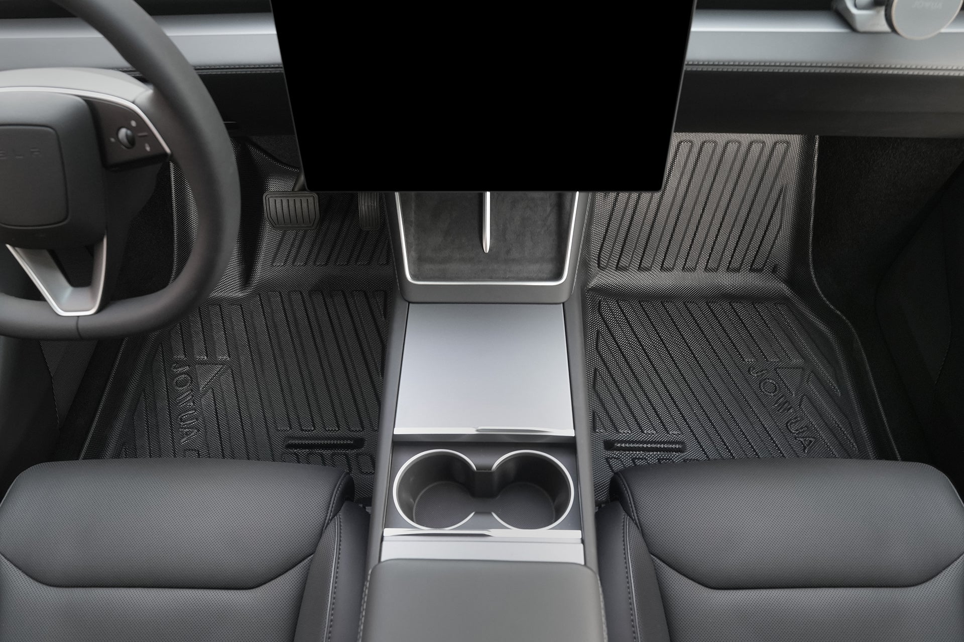 View of a car's center console with two cup holders, metallic accents, black leather seats, and JOWUA Model Y L (2026+) All Weather Floor Liners (Left Hand Drive). Air vent cover adds functionality. Steering wheel and pedals are partially visible.