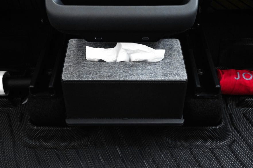 A gray tissue box with a textured cover sits on JOWUA All Model Y All-Weather Floor Liners Combo (Juniper 2025+ / 2021-2024) under a seat. Two tissues are partially pulled out, and a red item to the right adds to the accessory display.