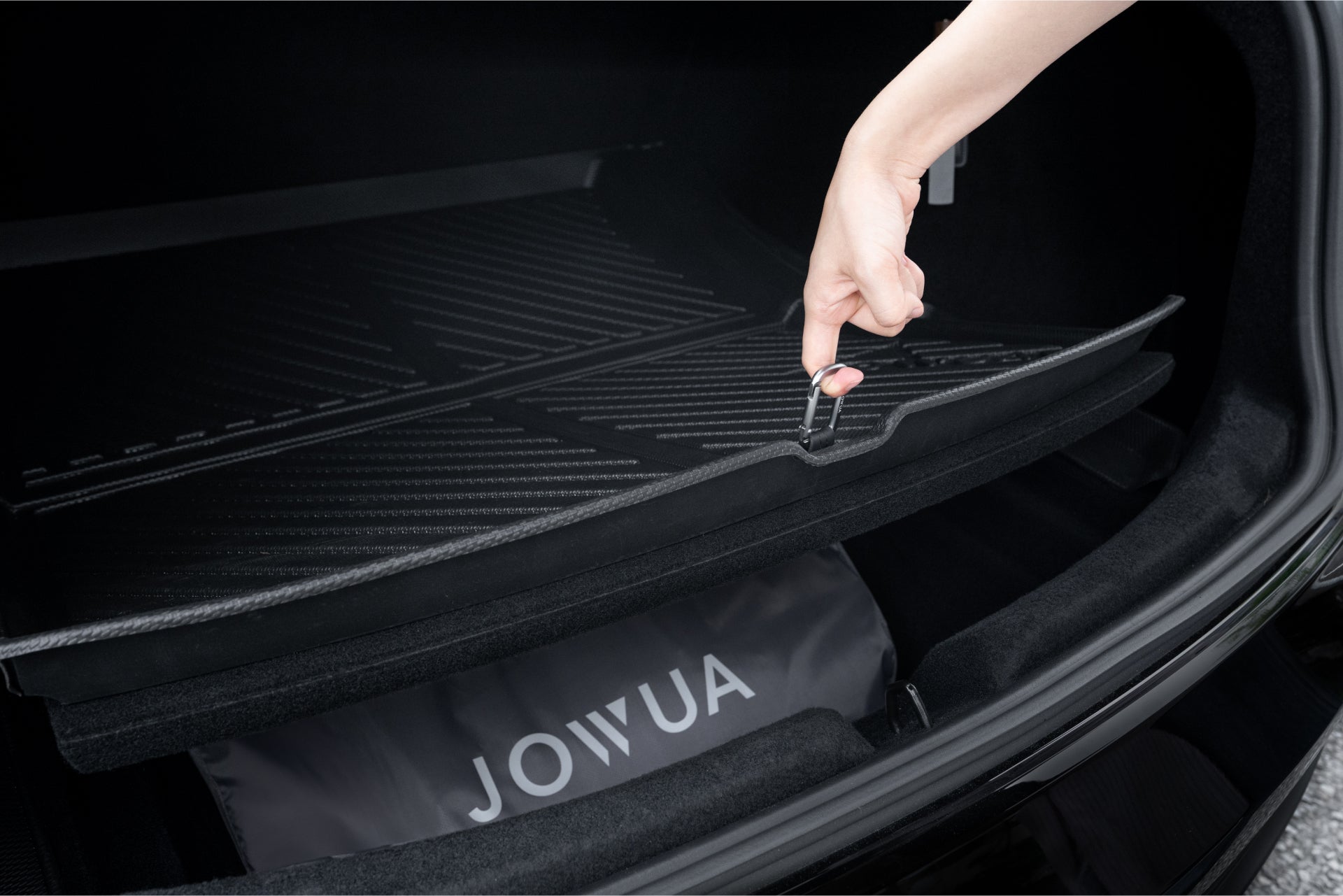 In the trunk of a Reengineered Model 3 Highland (2024+), someone lifts an all-weather floor mat featuring waterproof TPE material by a small handle. Underneath, a storage space is unveiled, revealing a bag marked with "JOWUA.