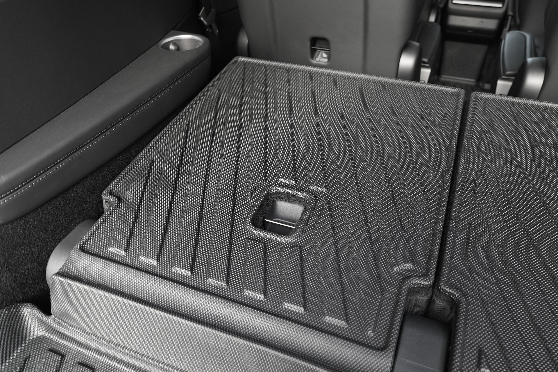 Close-up of folded rear seats with textured black upholstery and pull handle, forming a flat cargo area fitted with JOWUA Model Y L (2026+) Interior / Trunk Full Coverage Liner Combo; cupholders appear in the background.
