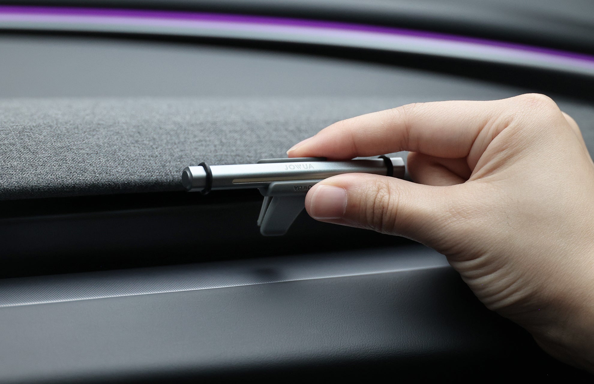 A hand holds a silver JOWUA Tesla Air Freshener for Model S/3/X/Y with a silicone base near a car's fabric dashboard, as a purple accent glows in the background.