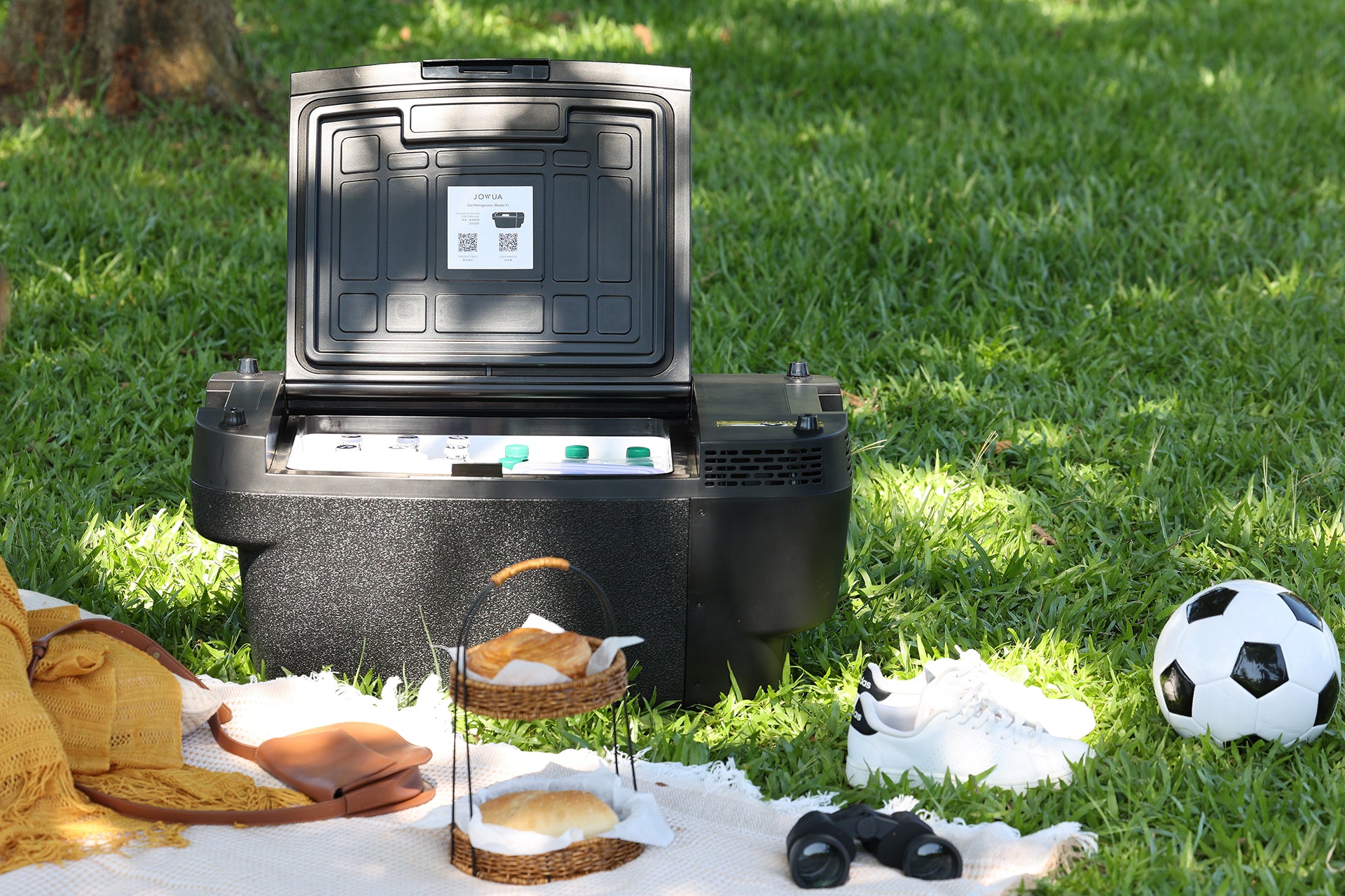 A JOWUA Car Refrigerator for Tesla Model Y with an open lid, featuring intelligent dual-mode cooling, sits on the grass beside a picnic setup with bread, a blanket, shoes, binoculars, and a soccer ball on a sunny day.
