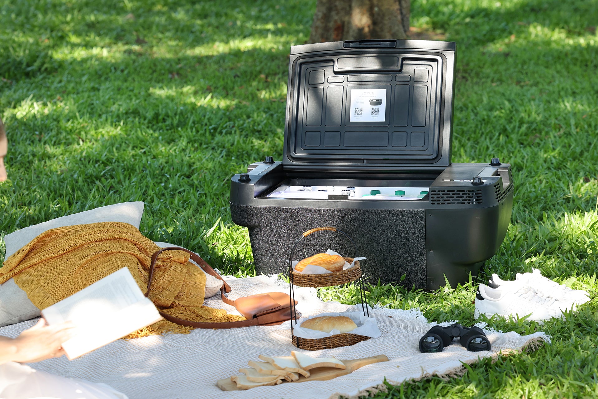 A JOWUA Car Refrigerator for Tesla Model Y with intelligent dual-mode cooling sits open on a picnic blanket on green grass, surrounded by food, drinks, shoes, headphones, and a person reading a book.