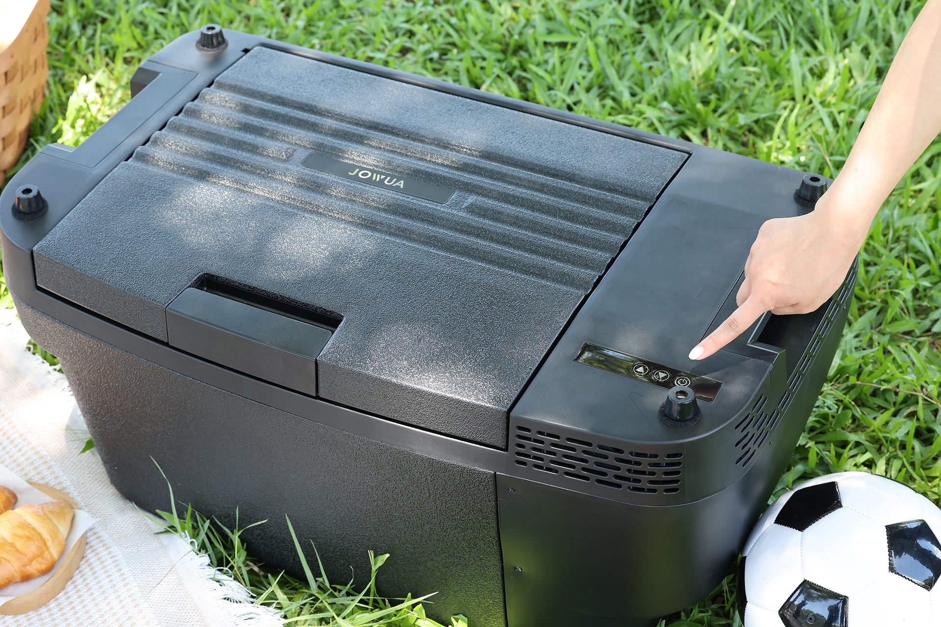 A person presses a button on the JOWUA Car Refrigerator for Tesla Model Y, a black portable cooler with intelligent dual-mode cooling, placed on grass near a soccer ball, croissants, and a picnic blanket.
