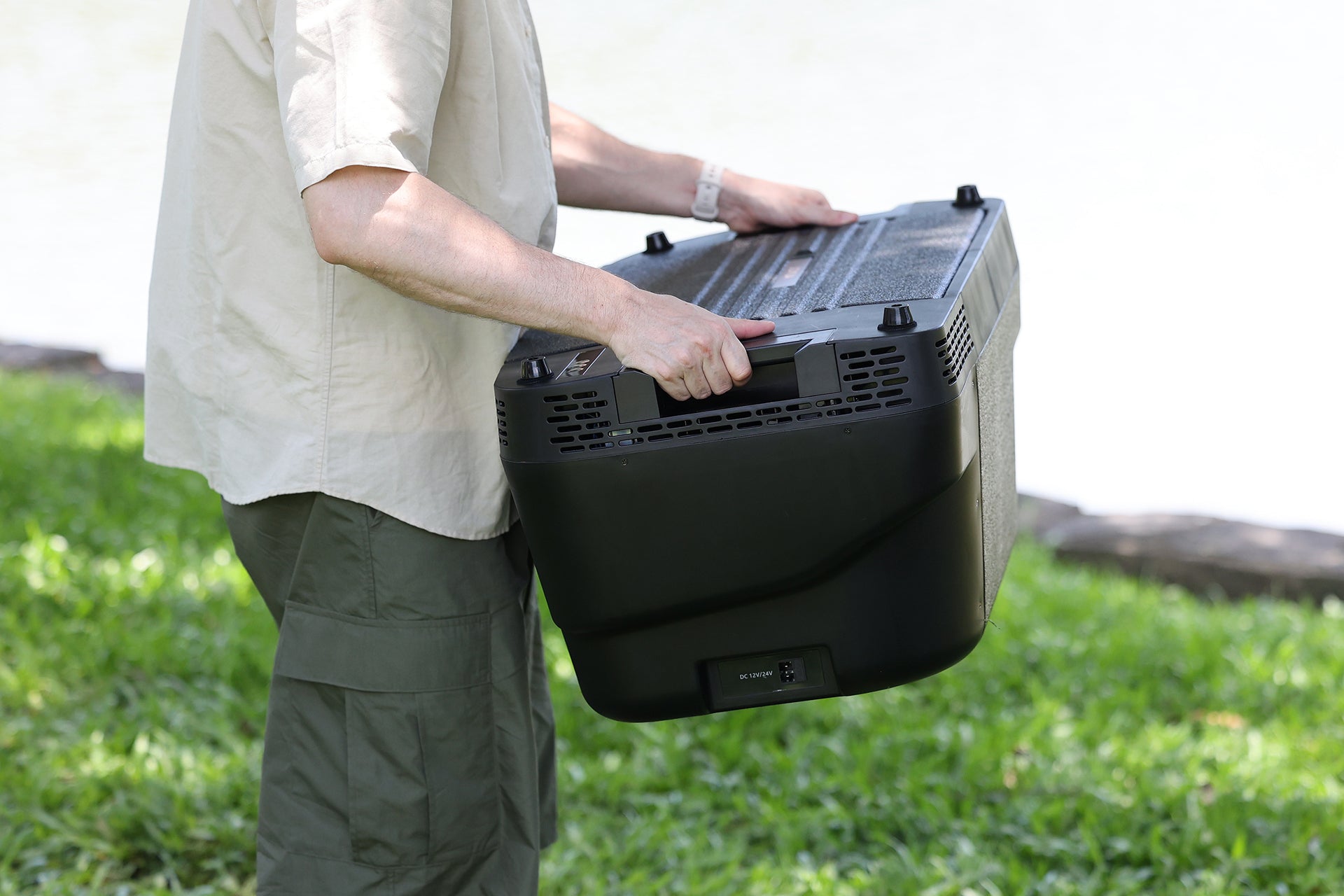 A person outdoors holds the JOWUA Car Refrigerator for Tesla Model Y, a large black portable appliance with handles and buttons, featuring an intelligent dual-mode cooling system, while standing on grass near the water.