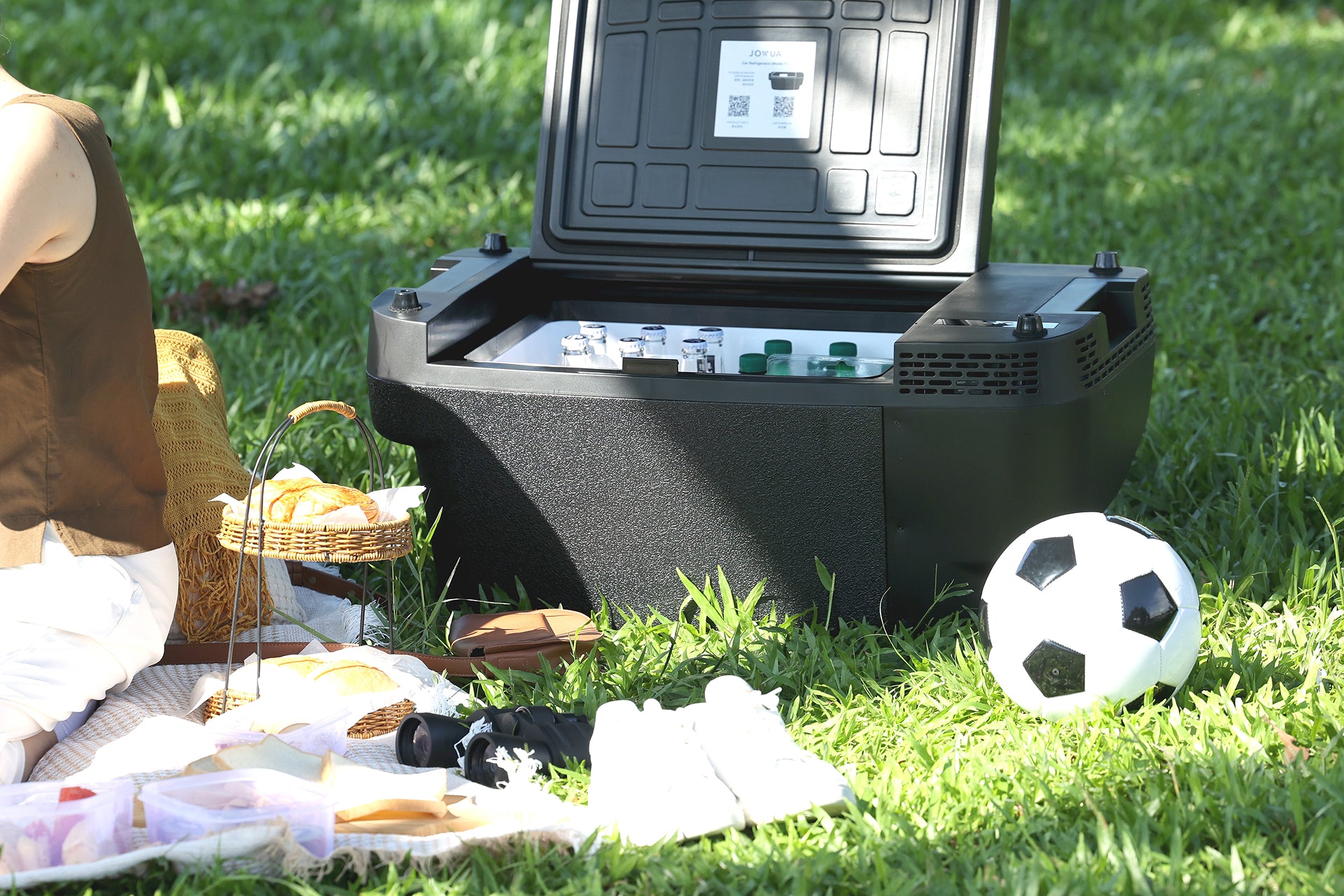A picnic scene on grass features a person sitting beside a blanket with a basket of food, white shoes, and a soccer ball, while drinks stay perfectly chilled in the JOWUA Car Refrigerator for Tesla Model Y with intelligent dual-mode cooling.