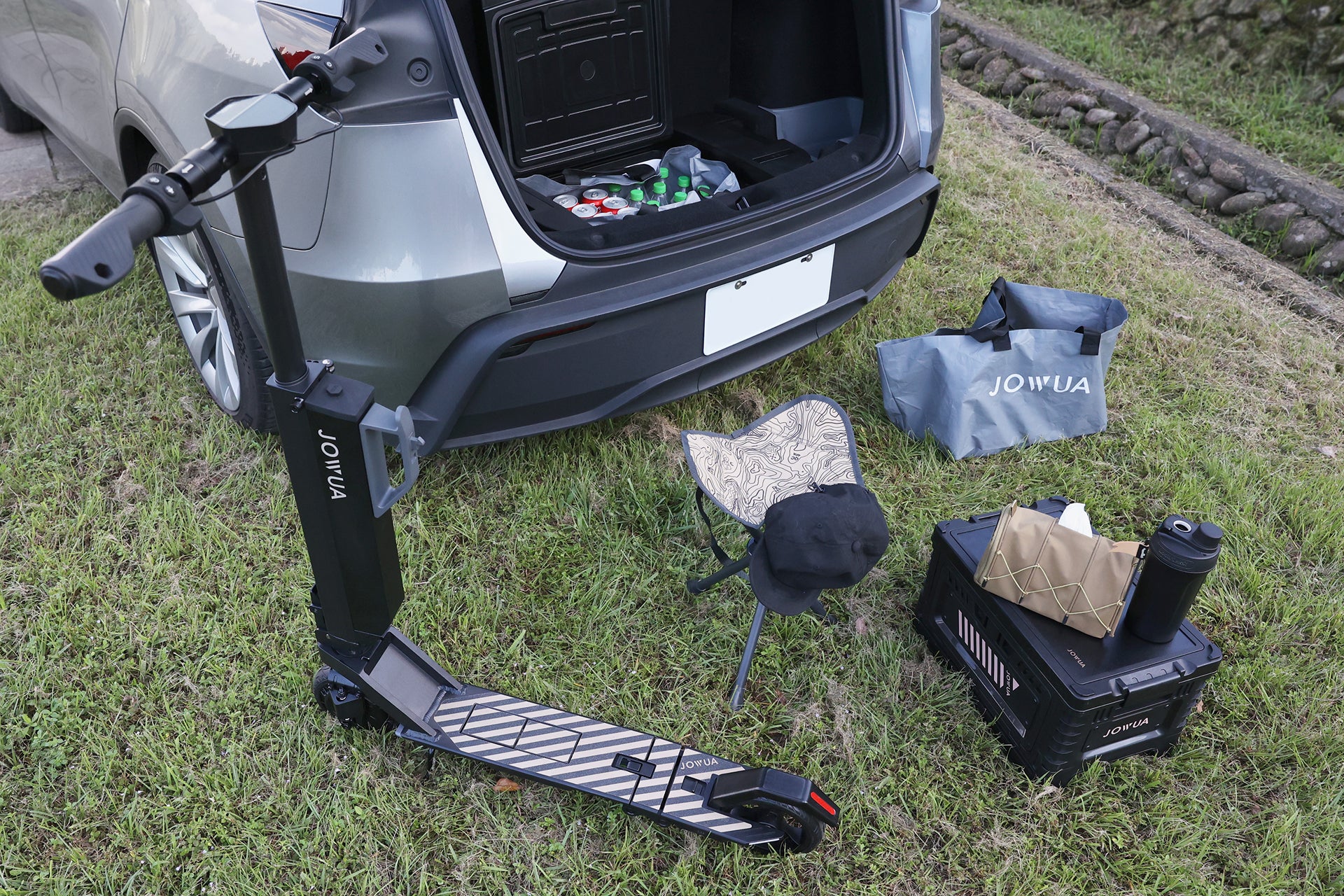 A car with its trunk open is parked on grass, featuring a JOWUA Car Refrigerator for Tesla Model Y among items like a black USB-C electric scooter, folding chair, crate, thermos, bag, and picnic supplies arranged nearby.