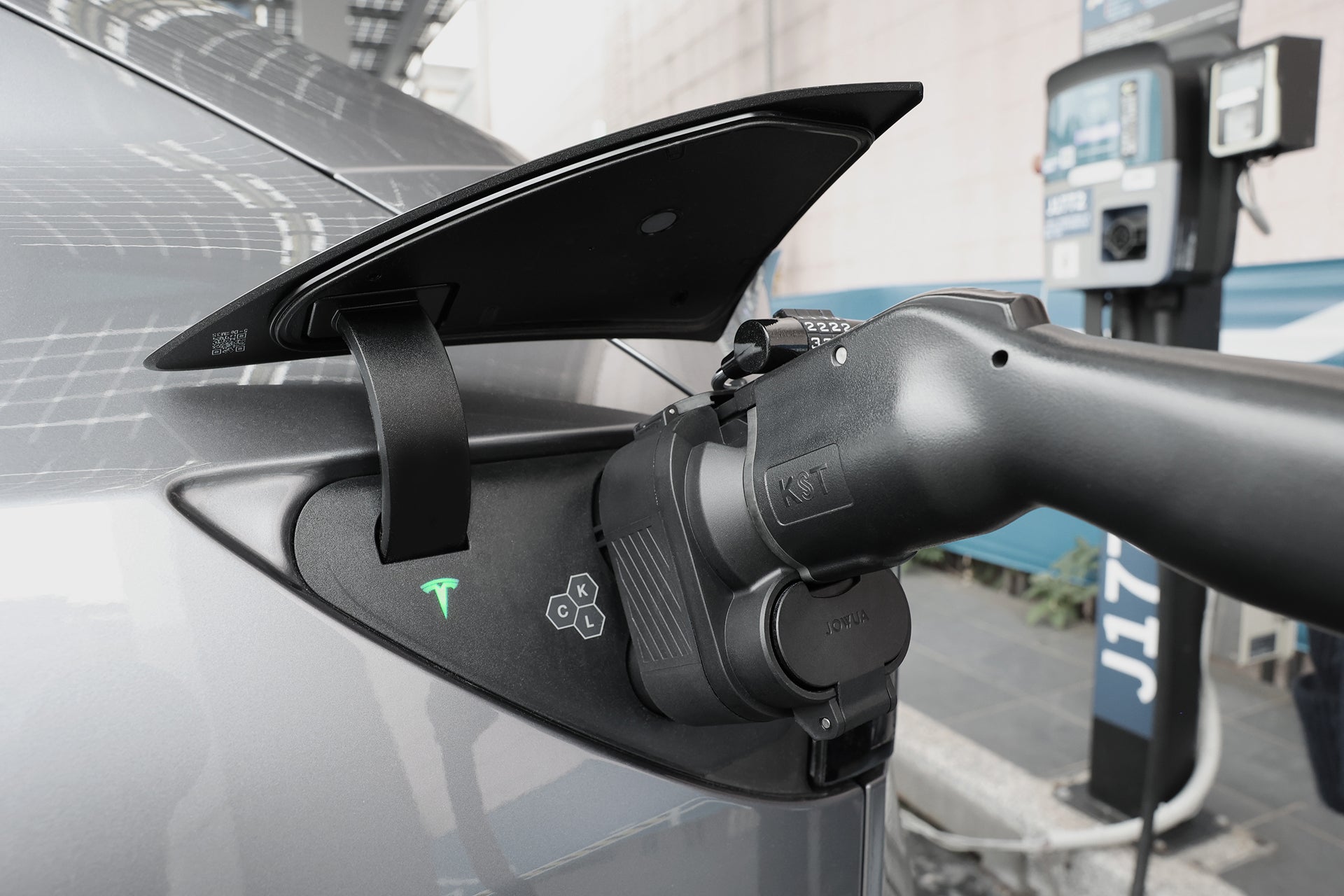 A close-up of a Tesla being charged at a station with the JOWUA CCS1 to CCS2 DC & AC Combined Adapter, showing the charging cable connected to the car’s port and the green Tesla logo illuminated.