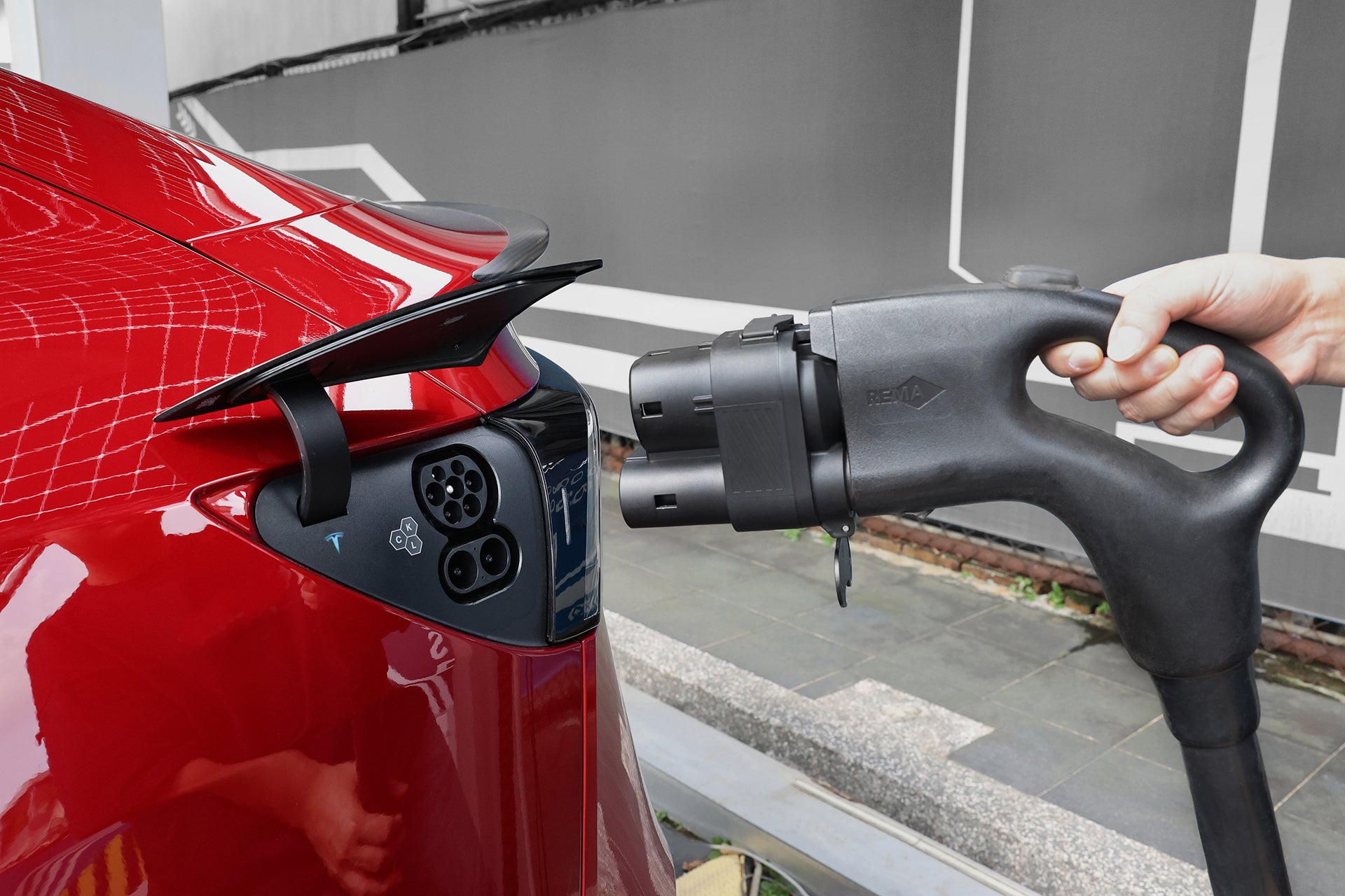 A person holds a JOWUA CCS1 to CCS2 DC & AC Combined Adapter electric vehicle charging plug near the charging port of a red car on a paved outdoor area with a gray wall in the background, ready to connect it.