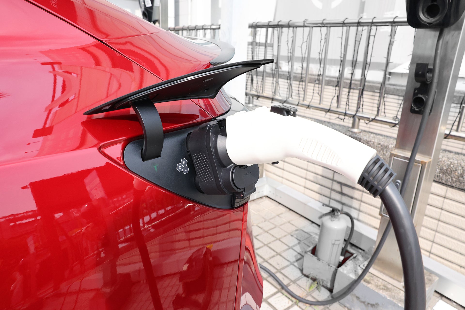 A close-up shows a red EV charging with the JOWUA CCS1 to CCS2 DC & AC Combined Adapter, plugged into the vehicle's port. The scene features a fence and pavement, emphasizing modern charging compatibility for electric vehicles.