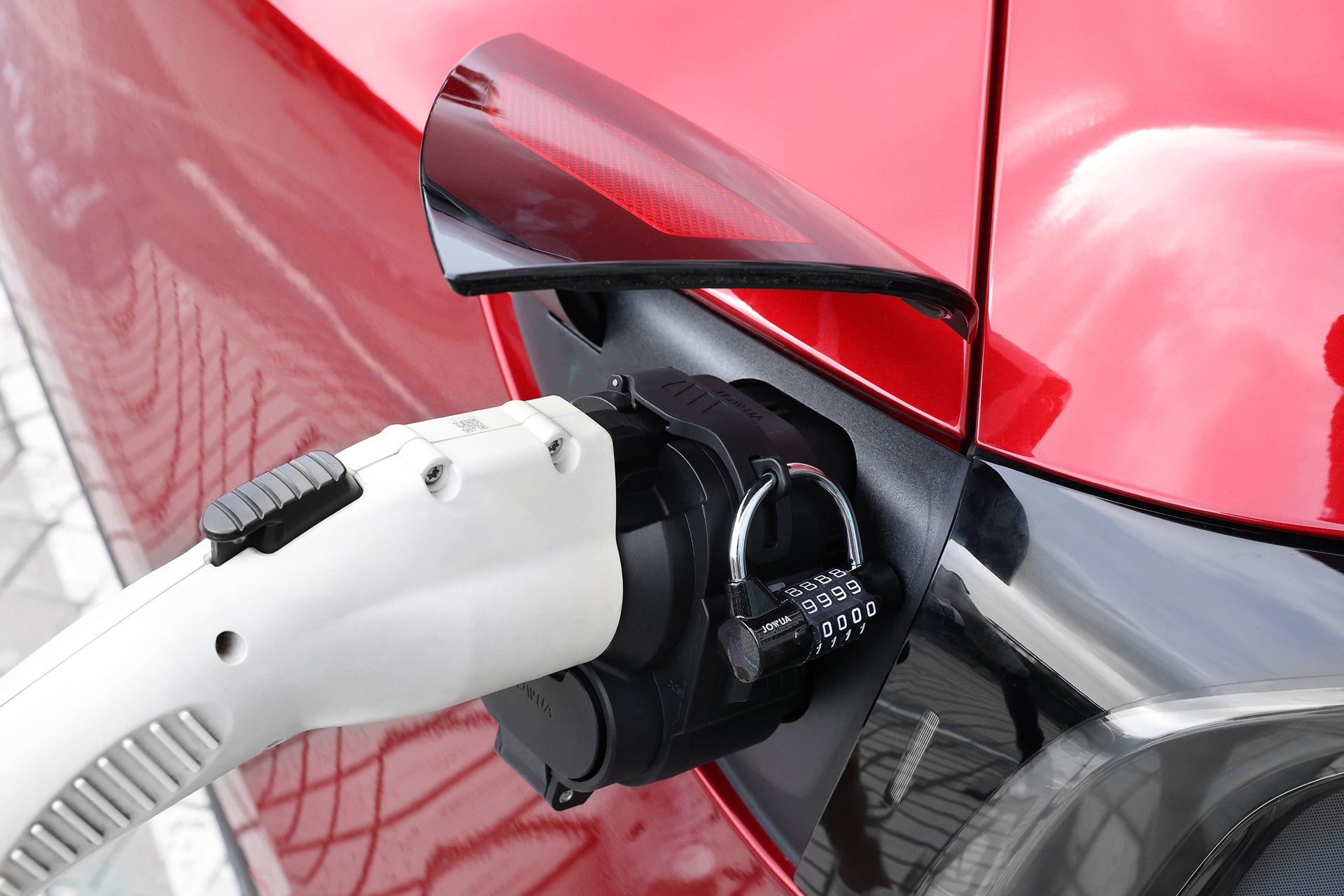 A white JOWUA CCS1 to CCS2 DC & AC Combined Adapter is plugged into a red EV’s charging port, secured with a combination padlock. The shiny car and pavement reflect compatibility for EV charging.
