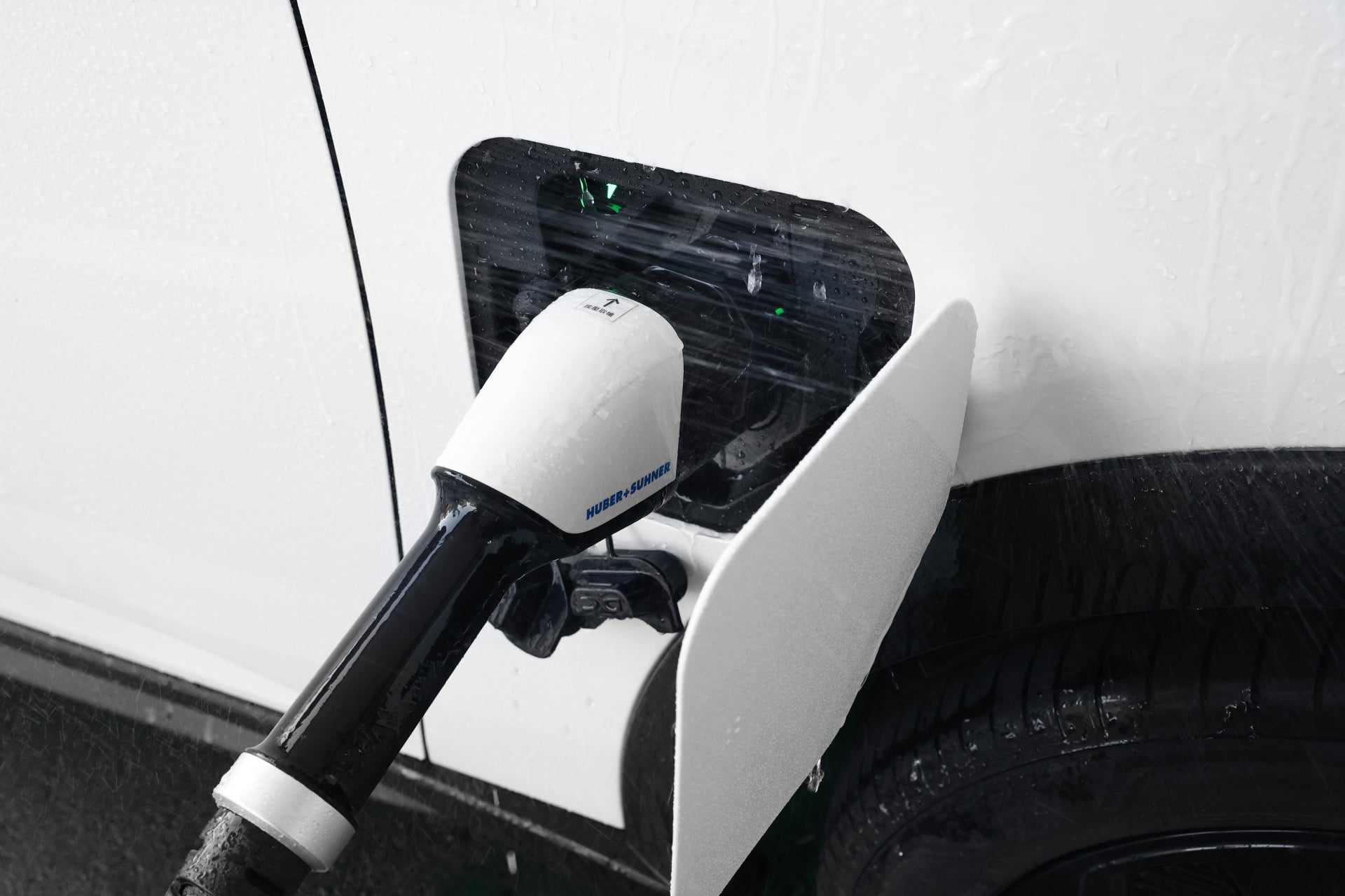 A white EV is charged using a JOWUA CCS2 to CCS1 DC & AC Combined Adapter, with the adapter plugged into the charging port. Water droplets on the open flap highlight its effective rapid charging in wet conditions.