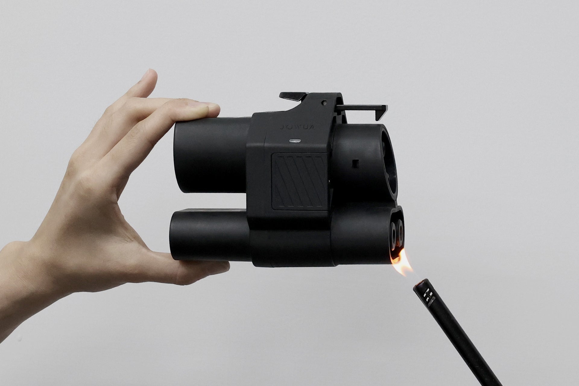 A hand holds a JOWUA CCS2 to CCS1 DC & AC Combined Adapter, featuring a black, boxy design with cylindrical barrels, while a lit lighter is placed near its lower part against a plain white background.