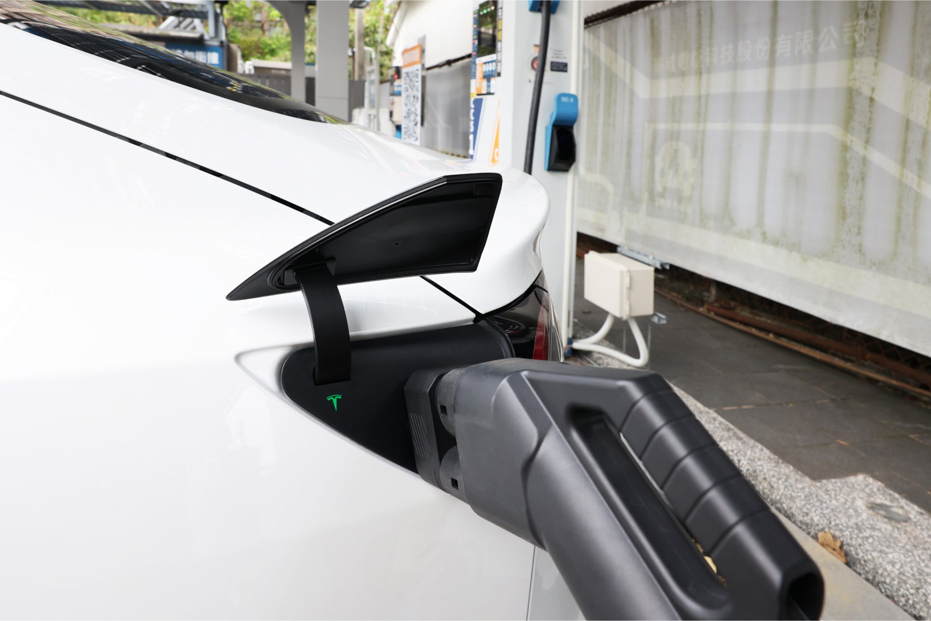 A white electric car is plugged into a charging station using a black JOWUA CCS2 to NACS DC Charging Adapter. The adapter smoothly fits the car