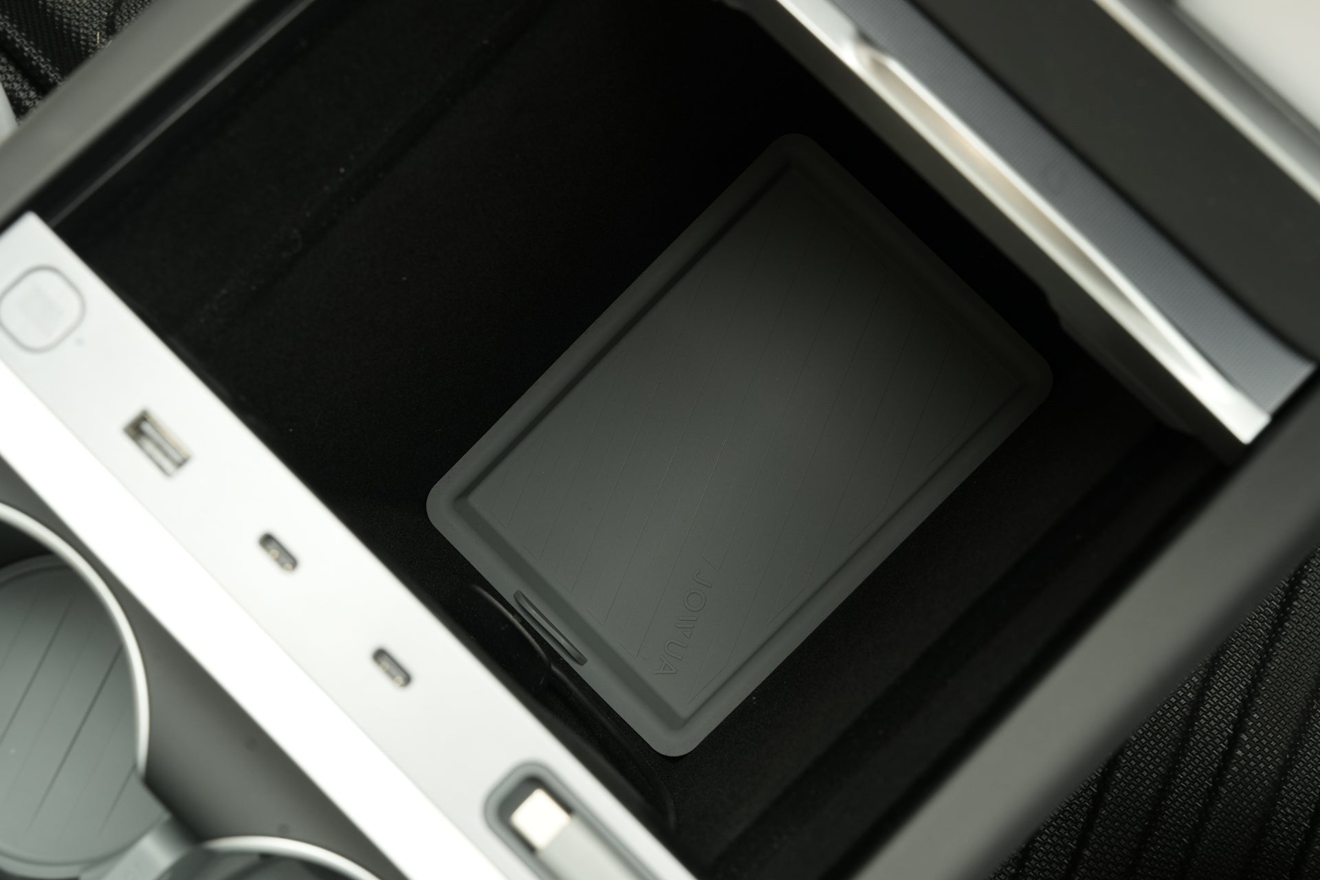 A gray tablet or e-reader sits at the bottom of a car’s center console storage, surrounded by cup holders and controls, featuring the JOWUA Center Console Set for Tesla Model Y Juniper 2025+—an essential accessory upgrade.