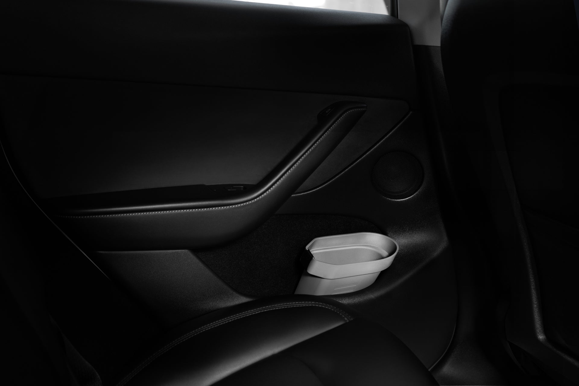 The rear seat area of a Tesla Model is highlighted by a sleek black door panel that includes a closed handle and an elegant small white storage bin from the JOWUA Door Side Storage Box set. The subtle lighting amplifies the minimalistic and modern design, with superior silicone material finishes enhancing its sophisticated appeal.