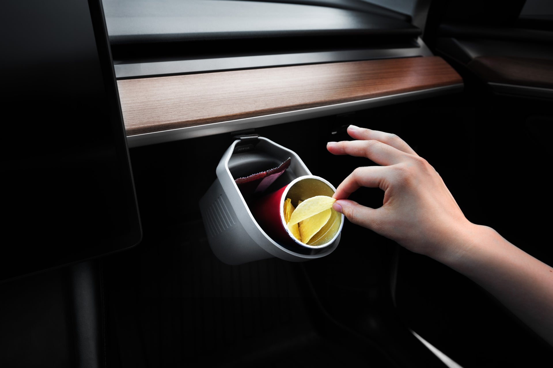 A hand places a single potato chip into a white cup filled with chips, nestled in a JOWUA Door Side Storage Box on the wooden dashboard of a Tesla Model S.