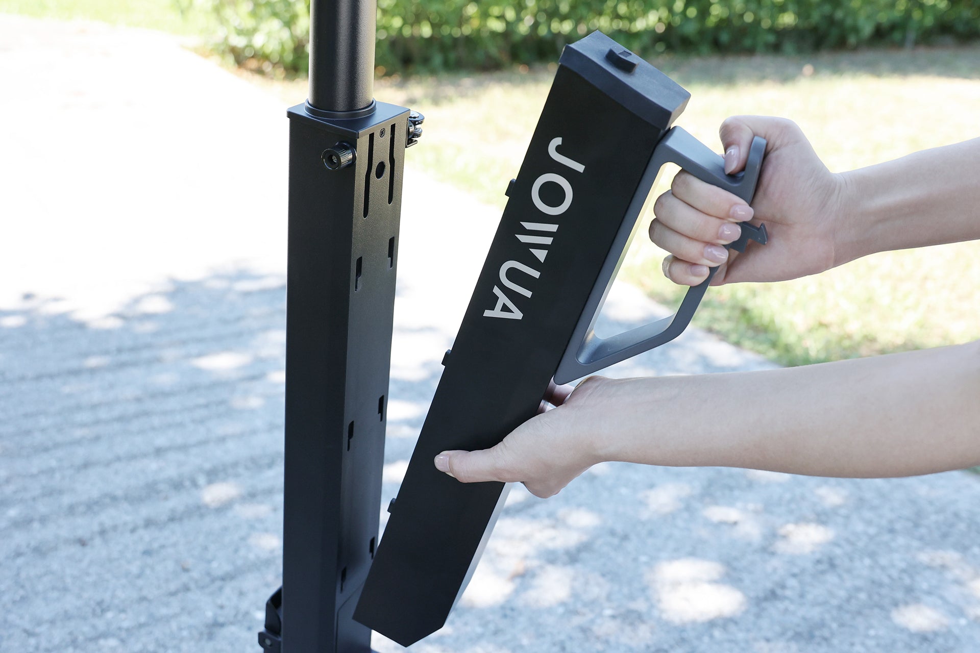 A person removes a rectangular black JOWUA battery from the vertical post of the Folding Electric Scooter｜Performance, with greenery and pavement in the background.