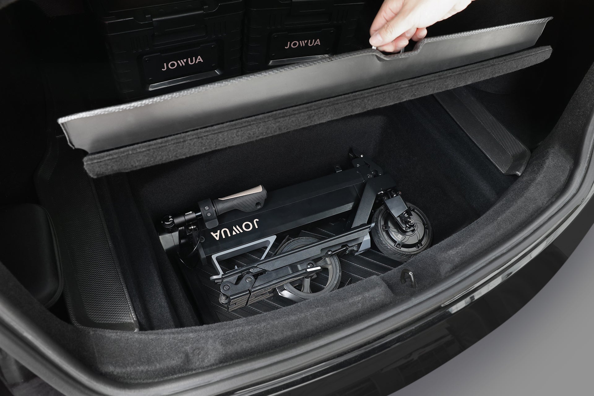 A hand lifts a car trunk cover, revealing a folded JOWUA Folding Electric Scooter｜Performance with long-range battery, neatly stored in the trunk compartment.