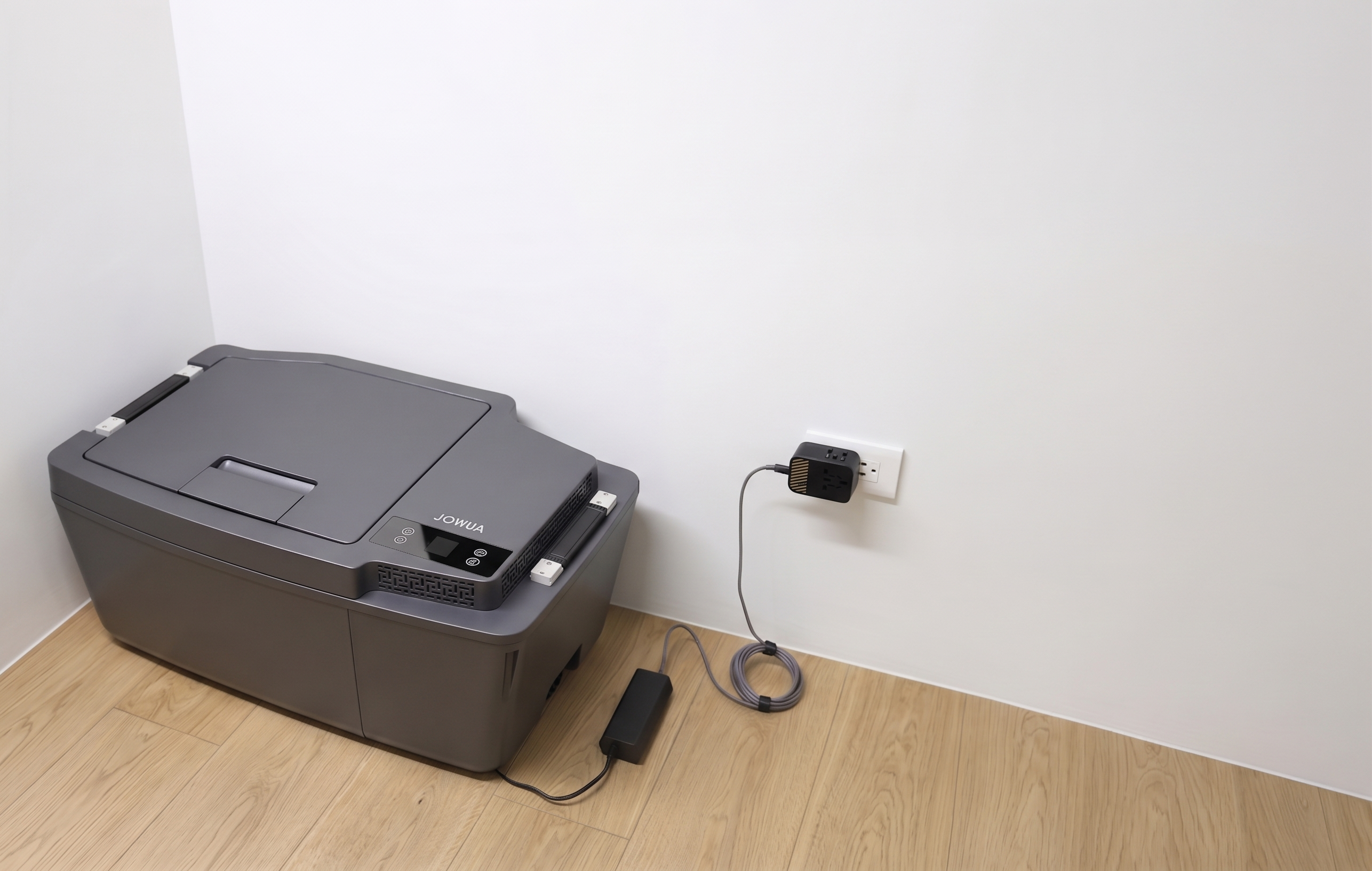 A JOWUA Car Refrigerator for Tesla Model 3 (Premium/Performance 2024-2026+) sits on a light wood floor near a white wall, plugged in with a USB-C power system. The area around the refrigerator and wall is uncluttered.