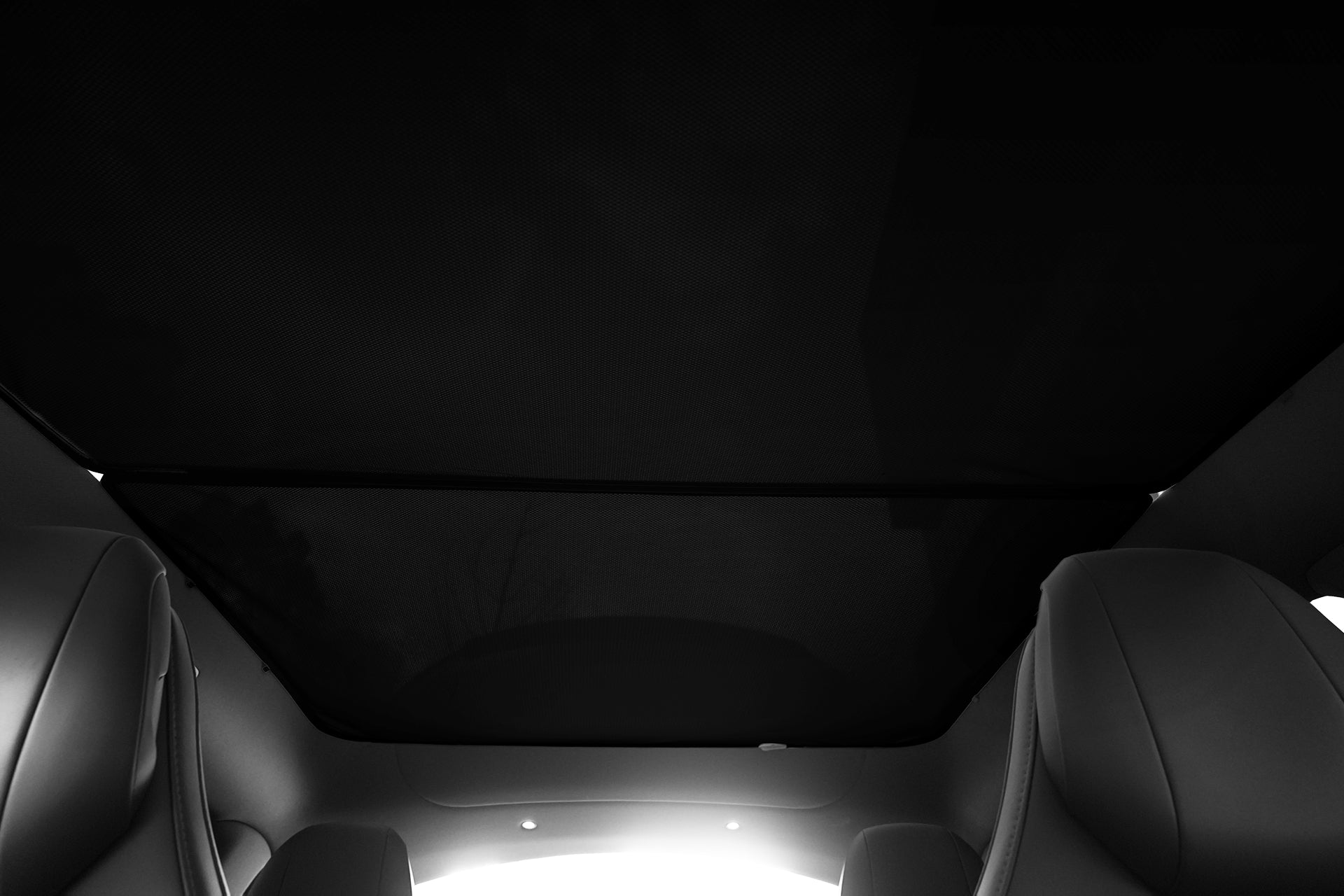 View from inside a car looking up at the JOWUA Model Y L (2026+) Glass Roof Sunshade with the sunshade closed; edges of two front seats visible, lighting highlights heat reduction and contrast between shaded and bright roof areas.