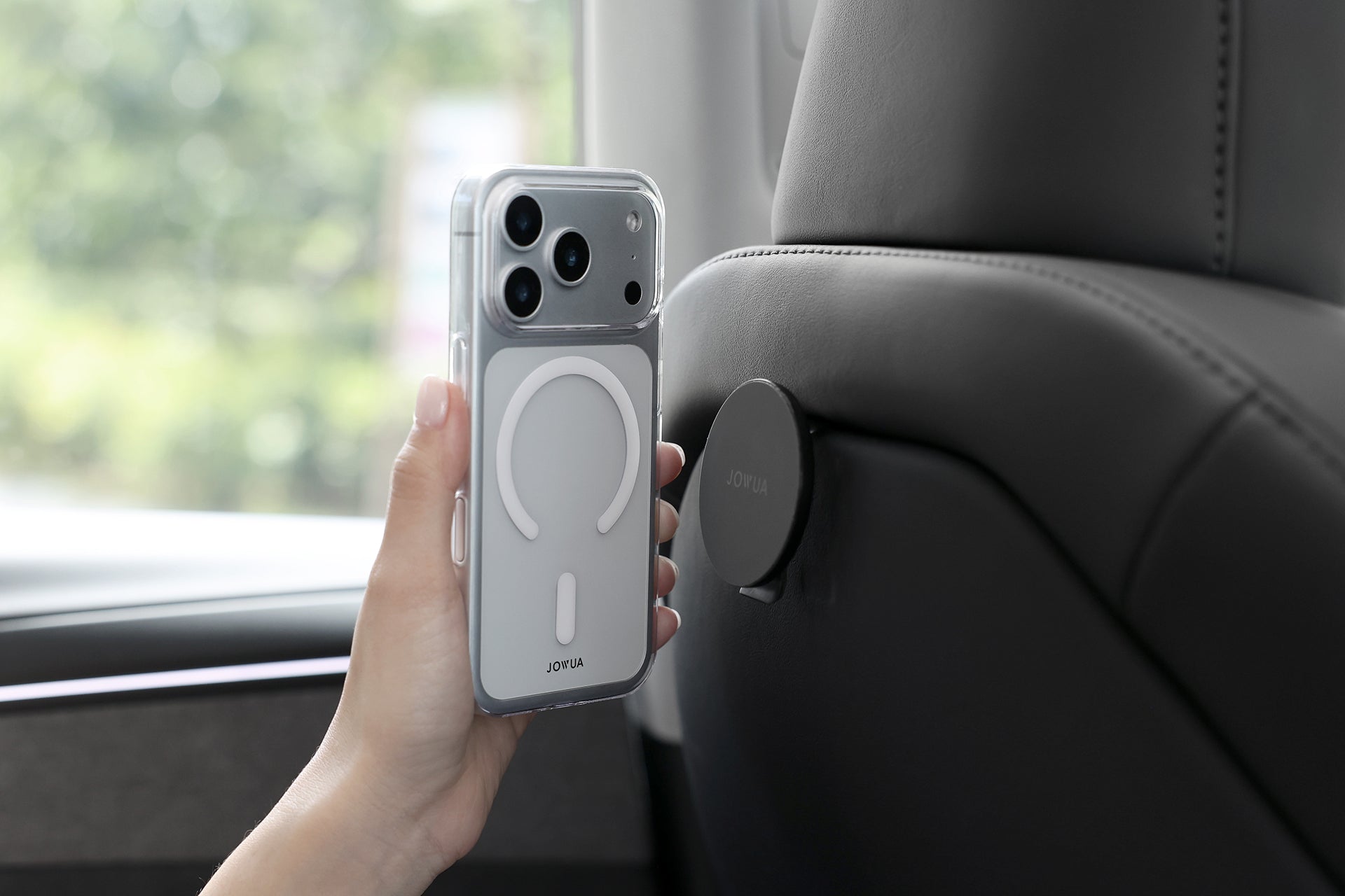 A hand holds a JOWUA iPhone Case for Tesla Car Mount with MagSafe + Phone Tether Tab Set (iPhone 17 Series) near a magnetic car mount on a headrest, with the car interior and blurred greenery in the background.